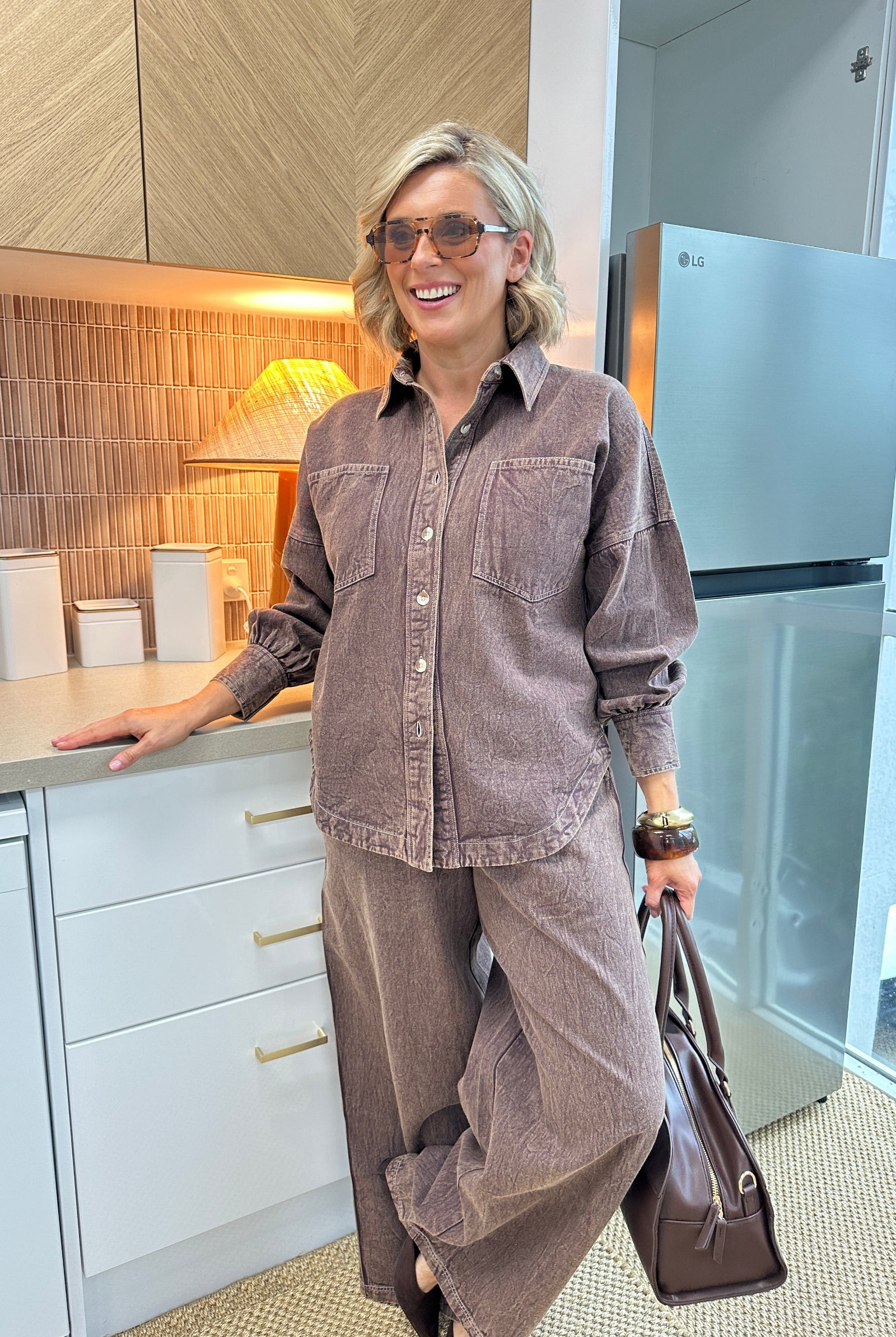 A woman wearing a matching brown corduroy button-up shirt and wide-leg trousers set, holding a brown leather bag in a modern kitchen setting.