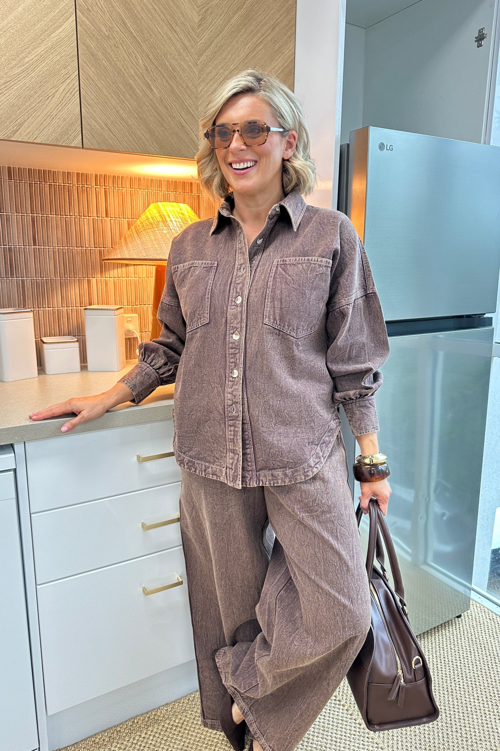 A woman wearing a matching brown corduroy button-up shirt and wide-leg trousers set, holding a brown leather bag in a modern kitchen setting.