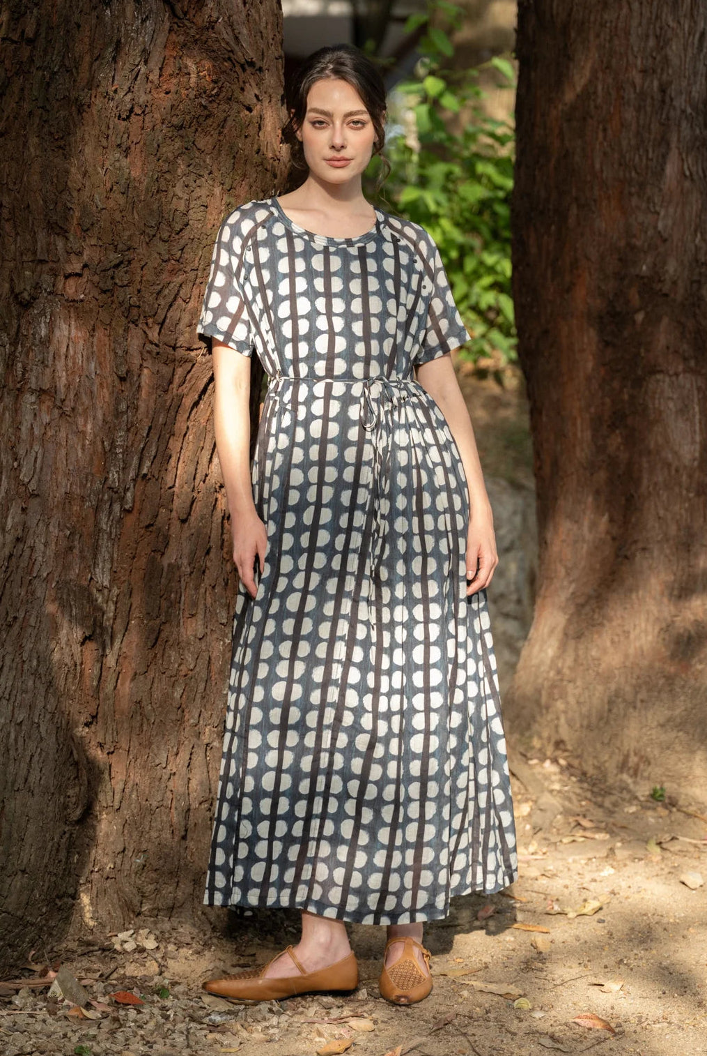A woman wearing a short-sleeve midi dress with a white and gray polka dot pattern, standing outdoors between two large tree trunks.