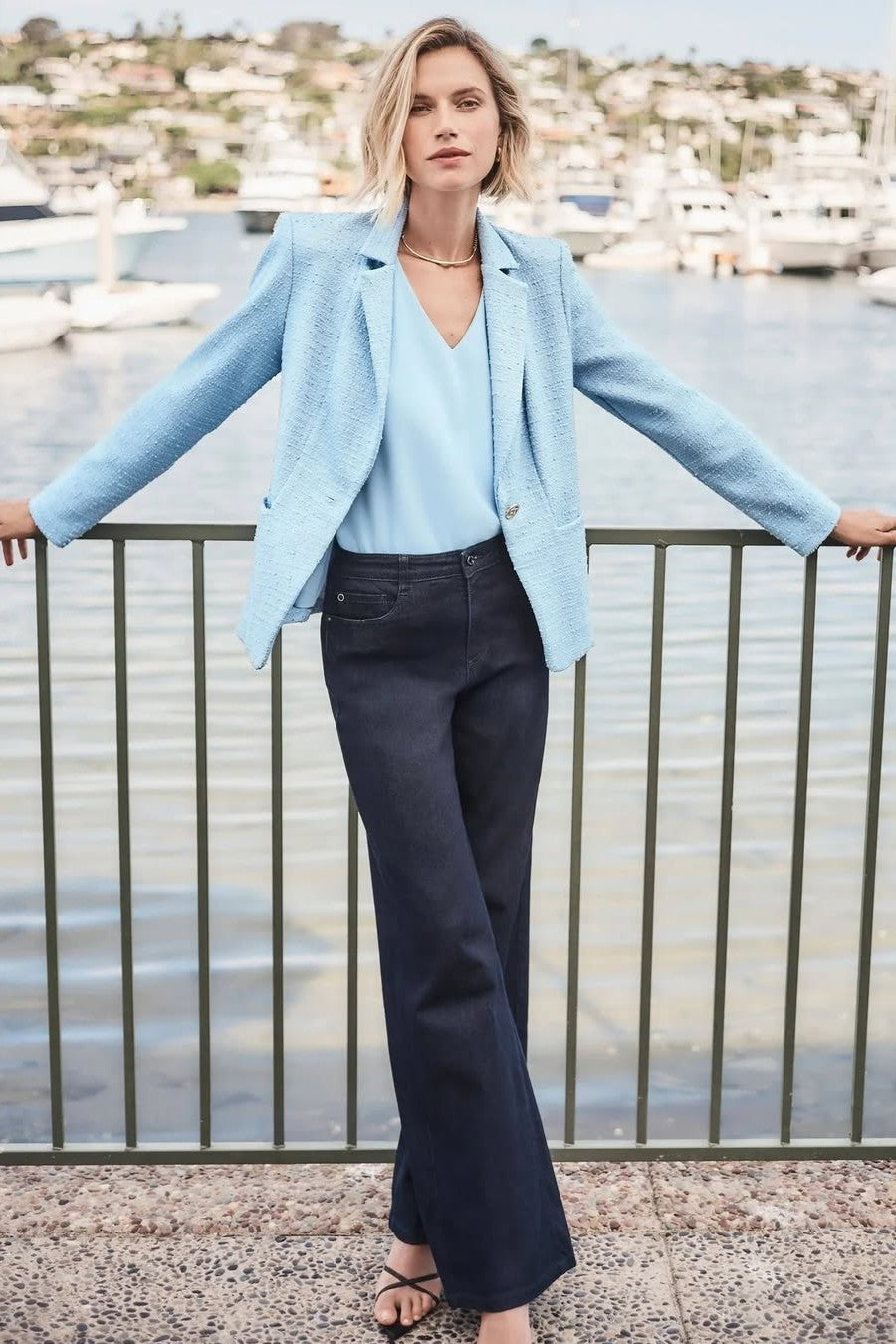 Woman in light blue blazer and dark pants standing by a waterfront with buildings in the background