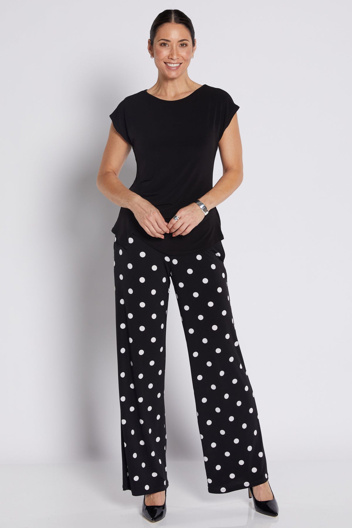 A woman modeling a black short-sleeve top and black wide-leg trousers with white polka dots, standing against a light gray studio background.