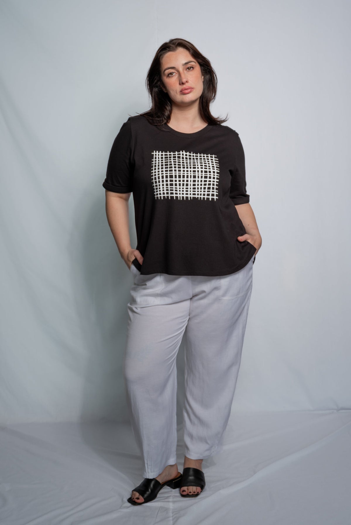 A woman modeling a black short-sleeve top with a white geometric patterned chest panel, paired with light gray wide-leg pants and black sandals, standing against a light gray studio backdrop.