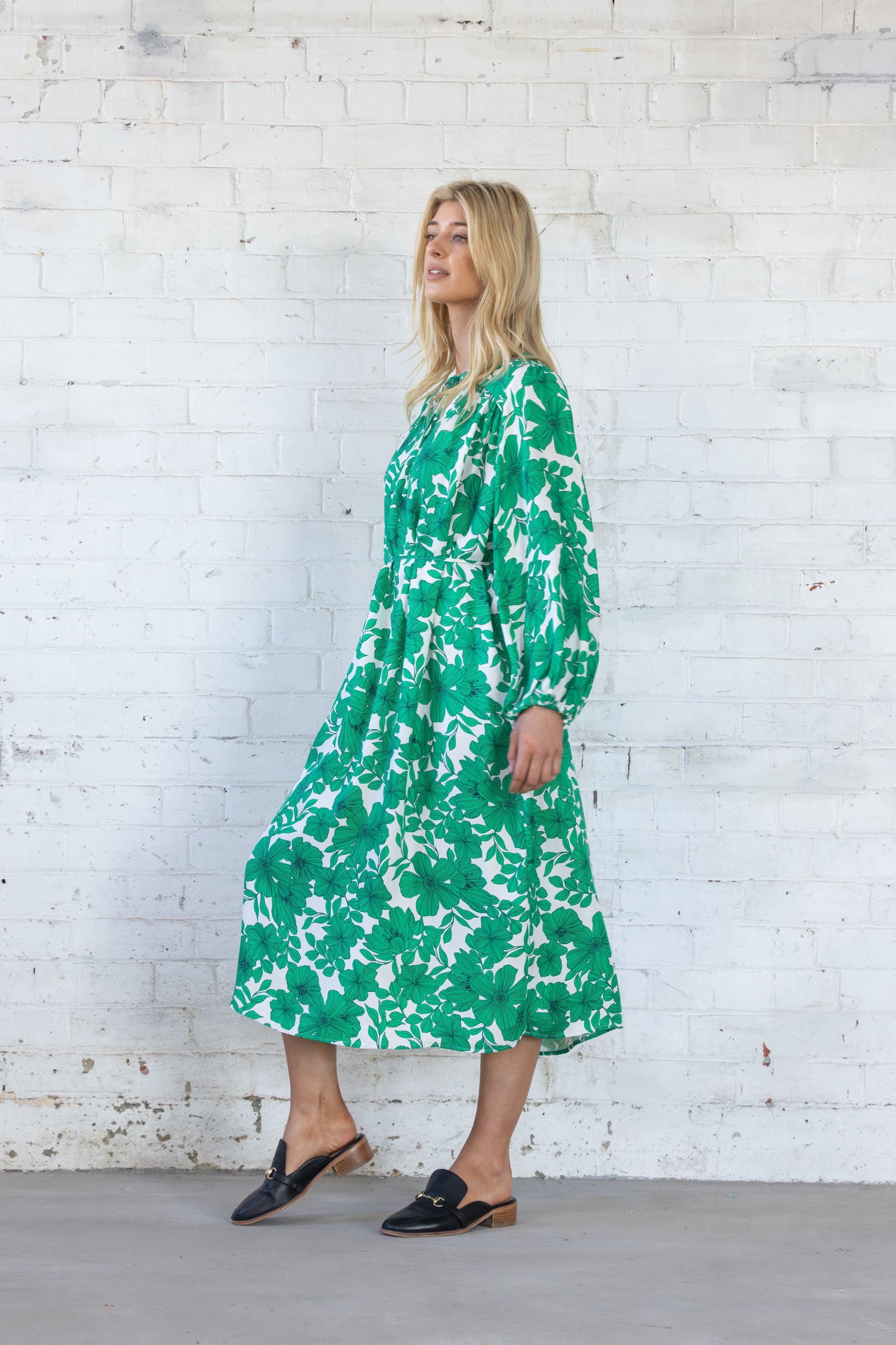 Woman wearing a green floral dress against a white brick wall.