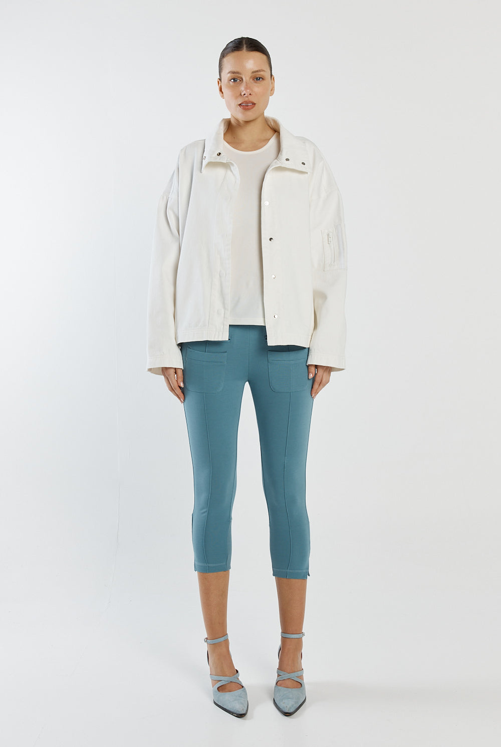 Woman wearing a white jacket, light blue shirt, and teal pants on a white background