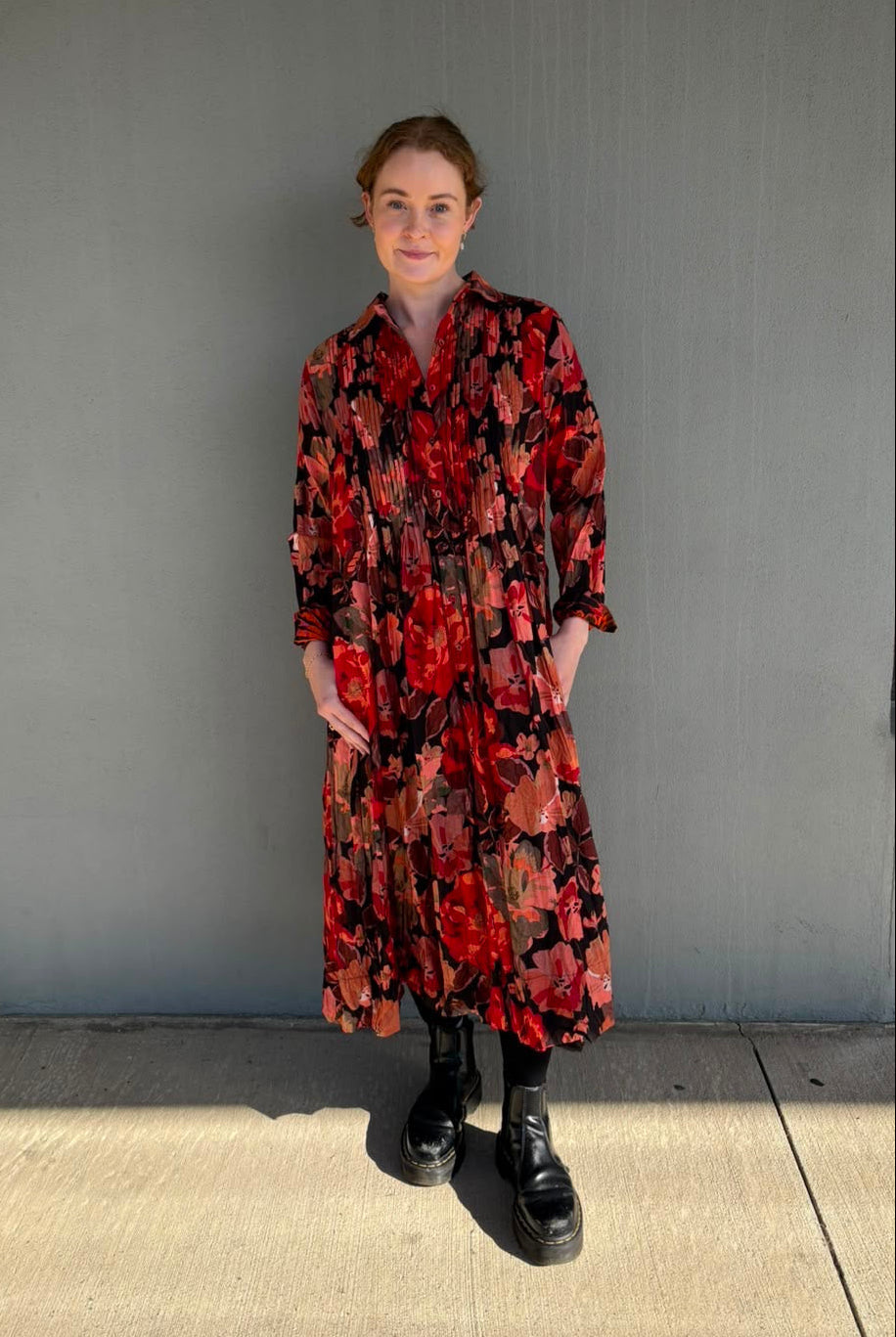 A woman stands outdoors against a gray building wall wearing a long-sleeve midi shirt dress with a bold red, black, and orange floral pattern, paired with black boots.