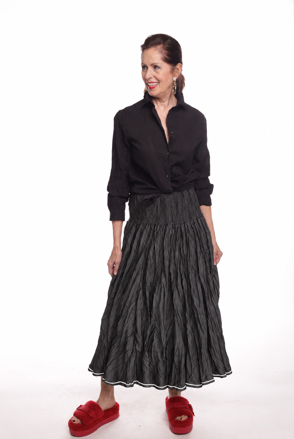 A woman modeling a black long-sleeve button-up shirt and a black pleated midi skirt, paired with red slippers, against a plain white background.