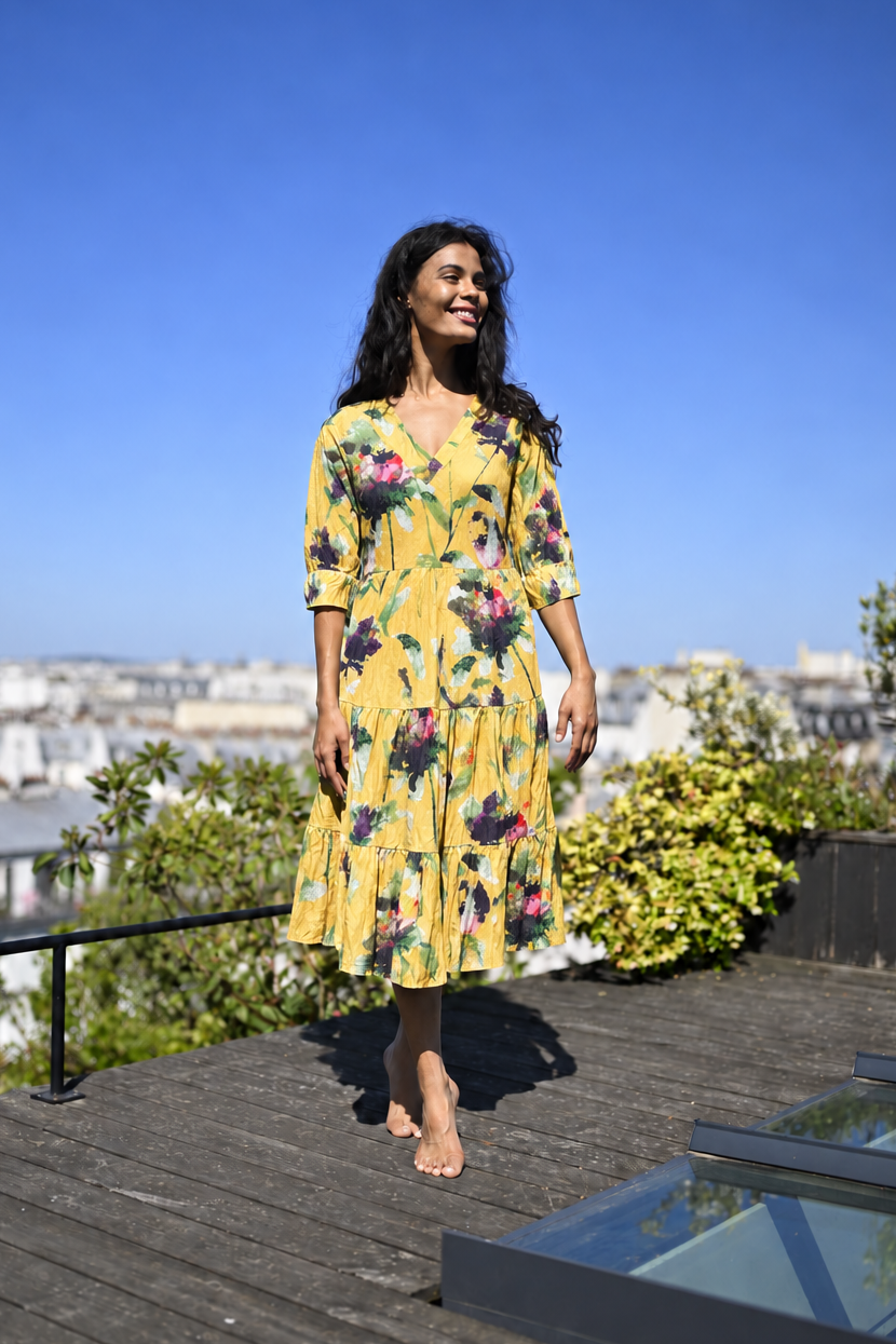 A woman wearing a yellow floral midi dress stands barefoot on a wooden deck with a city skyline and blue sky in the background.