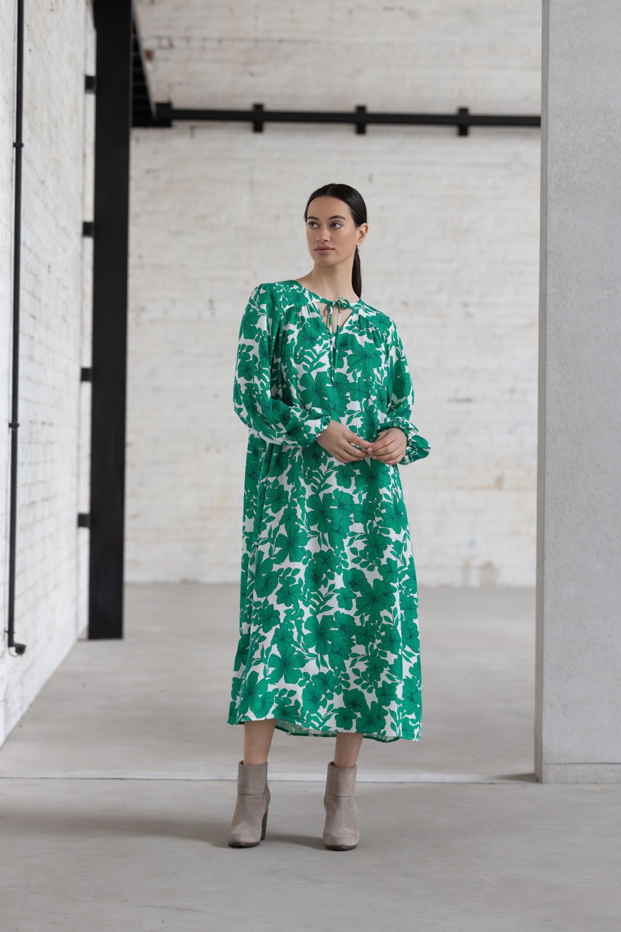 Woman wearing a green floral dress standing in a minimalistic room with white brick walls.