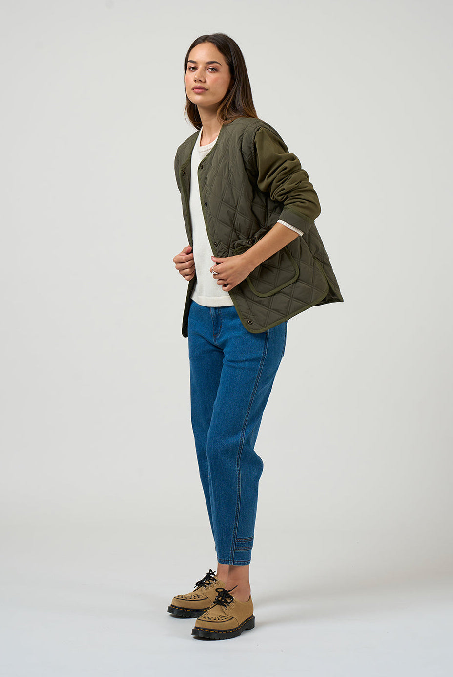 Woman wearing an olive green jacket, white shirt, blue jeans, and brown boots on a white background
