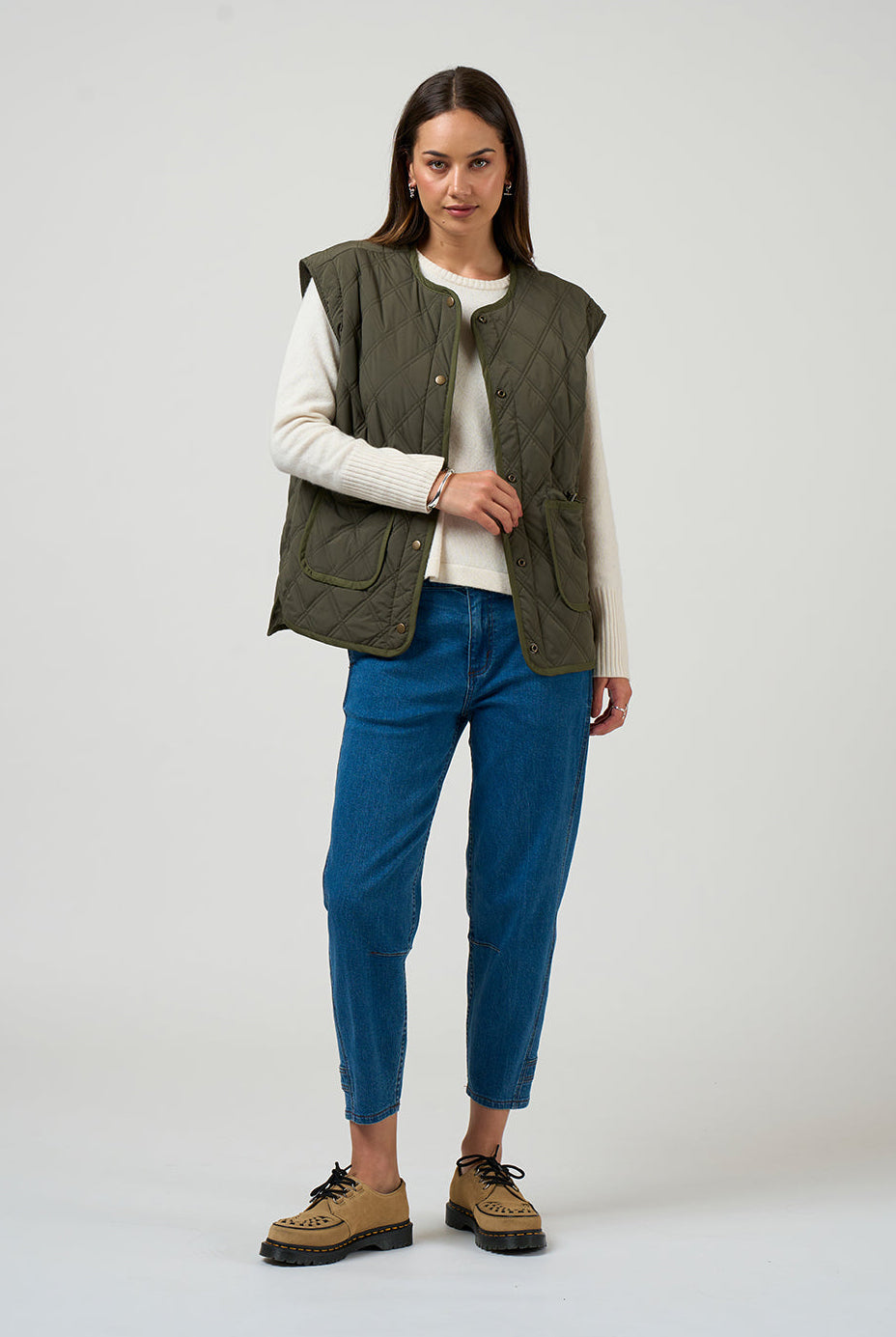 Woman wearing a green vest, white shirt, blue jeans, and brown shoes on a white background