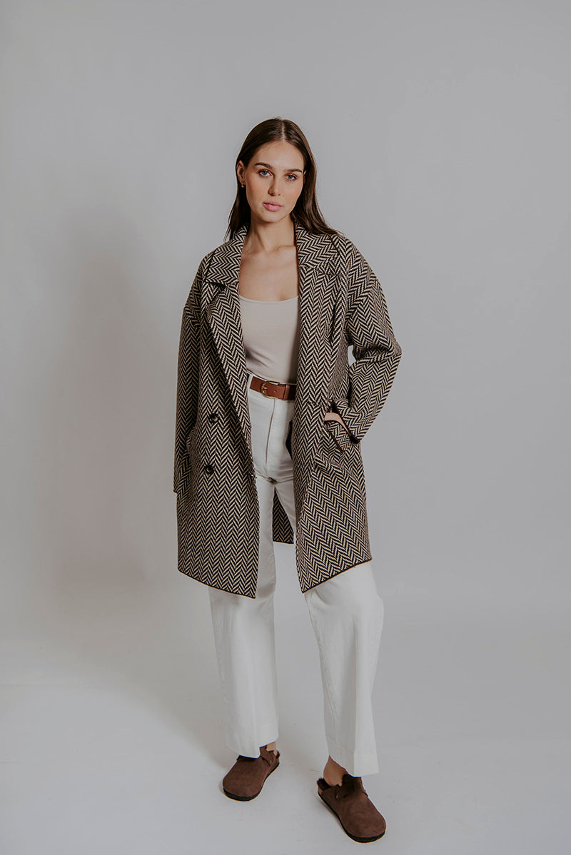 Woman wearing a long brown coat over a beige top and white pants on a plain background