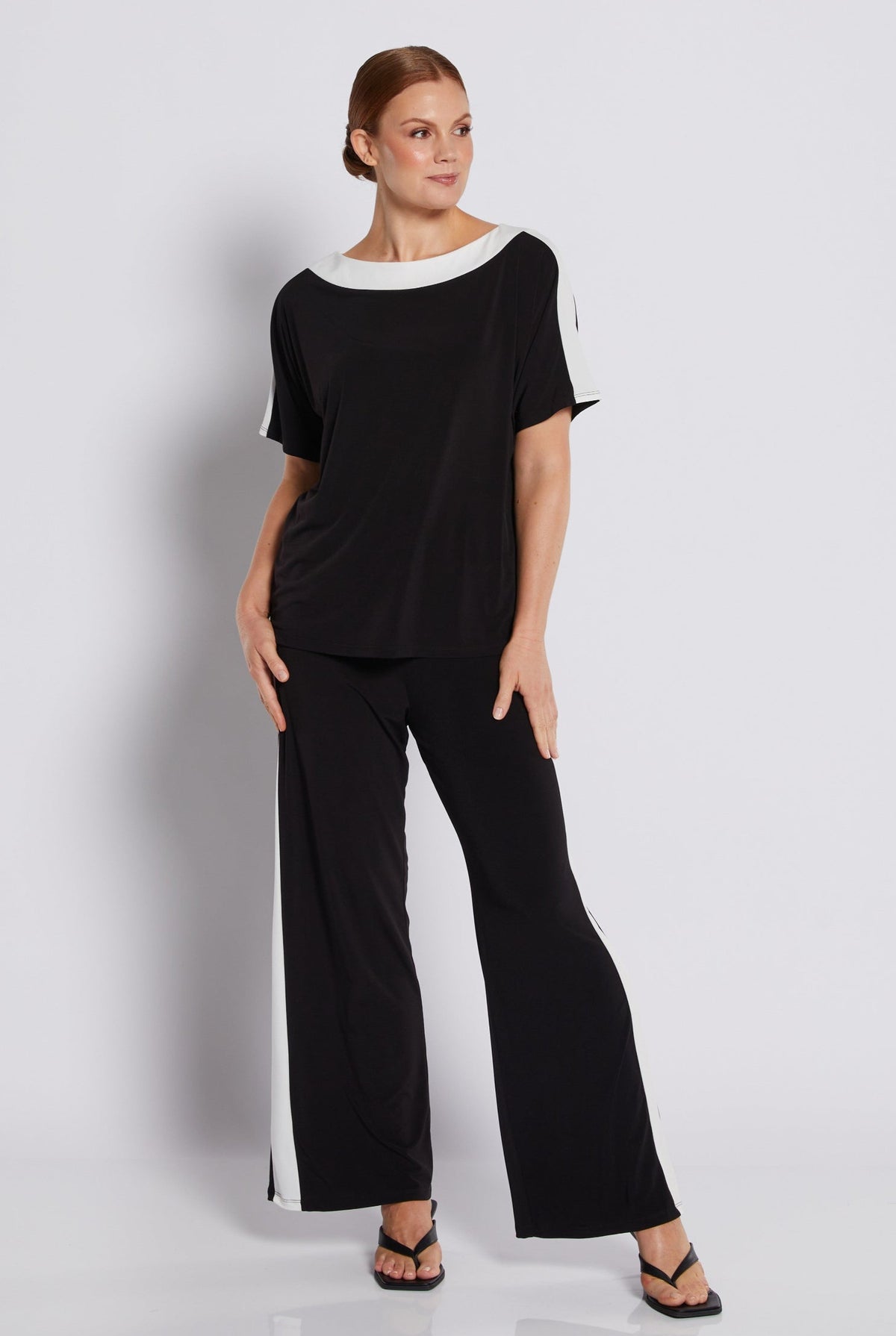 A woman modeling a black two-piece outfit consisting of a short-sleeve top with white contrast trim at the neckline and sleeve cuffs, and matching black wide-leg pants with white side panels, against a light gray studio background.
