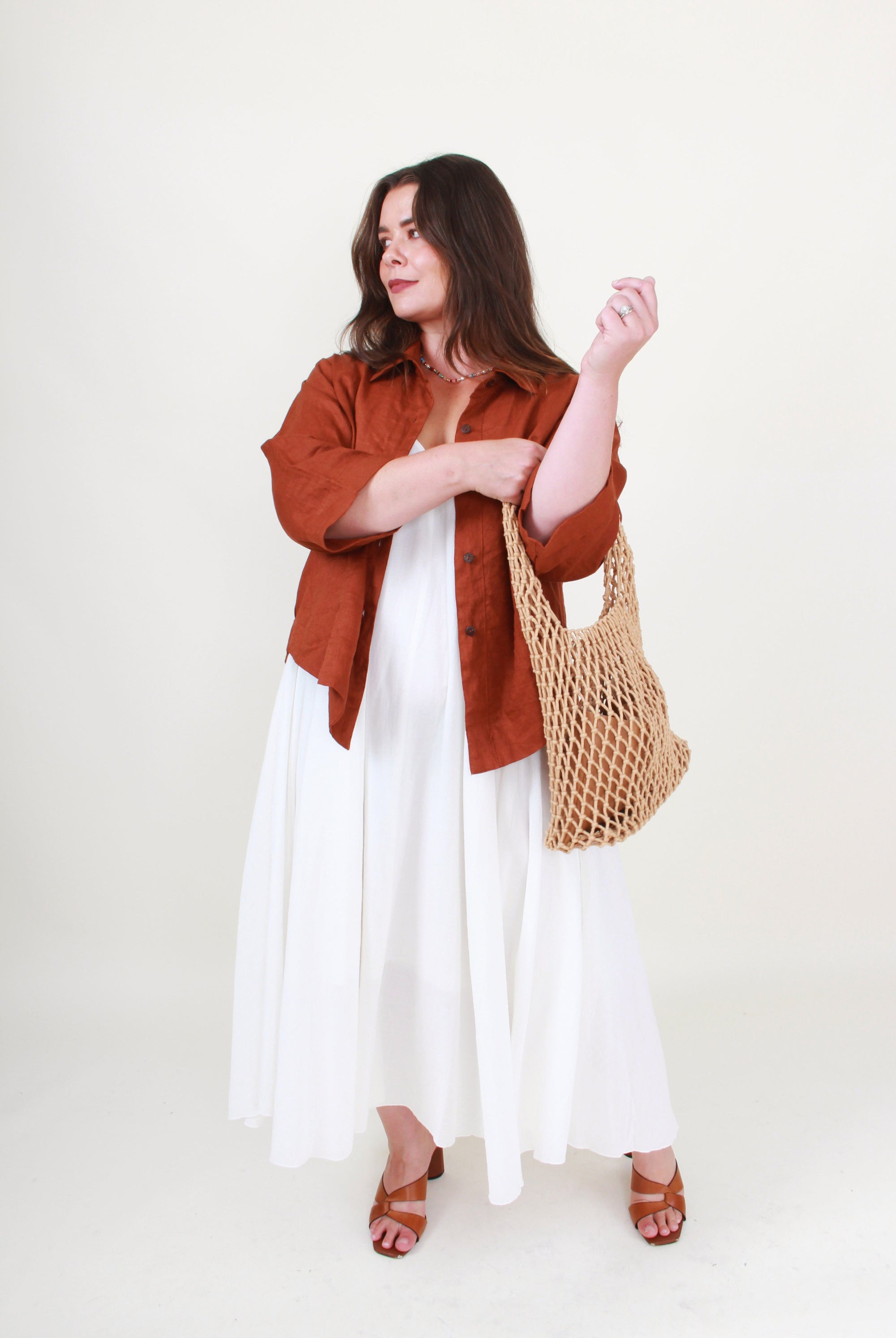 Woman wearing a rust-colored shirt, white dress, and holding a woven bag on a white background