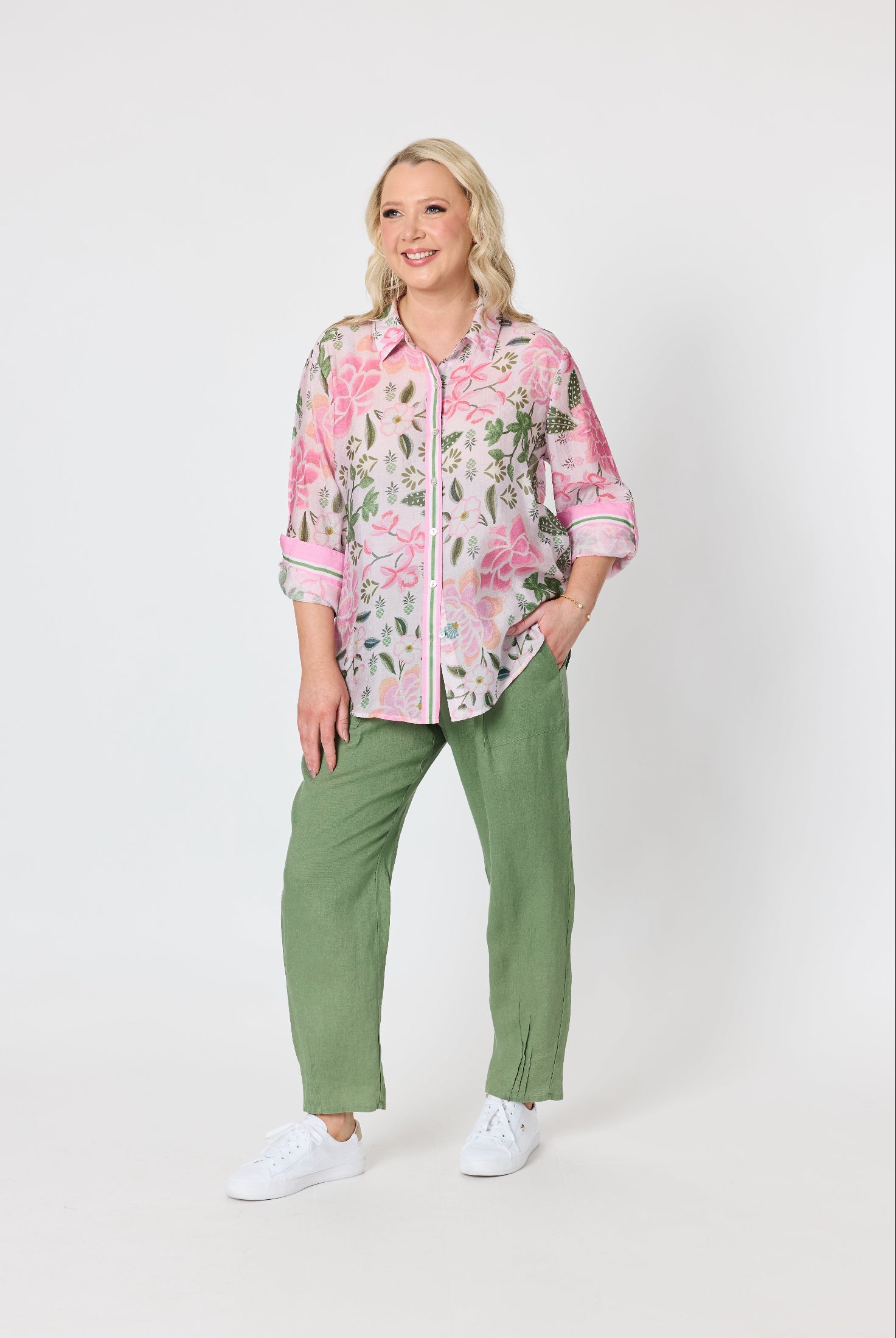 A woman modeling a pink and green floral button-up shirt with rolled sleeves, paired with olive green pants and white sneakers, against a plain light gray studio background.