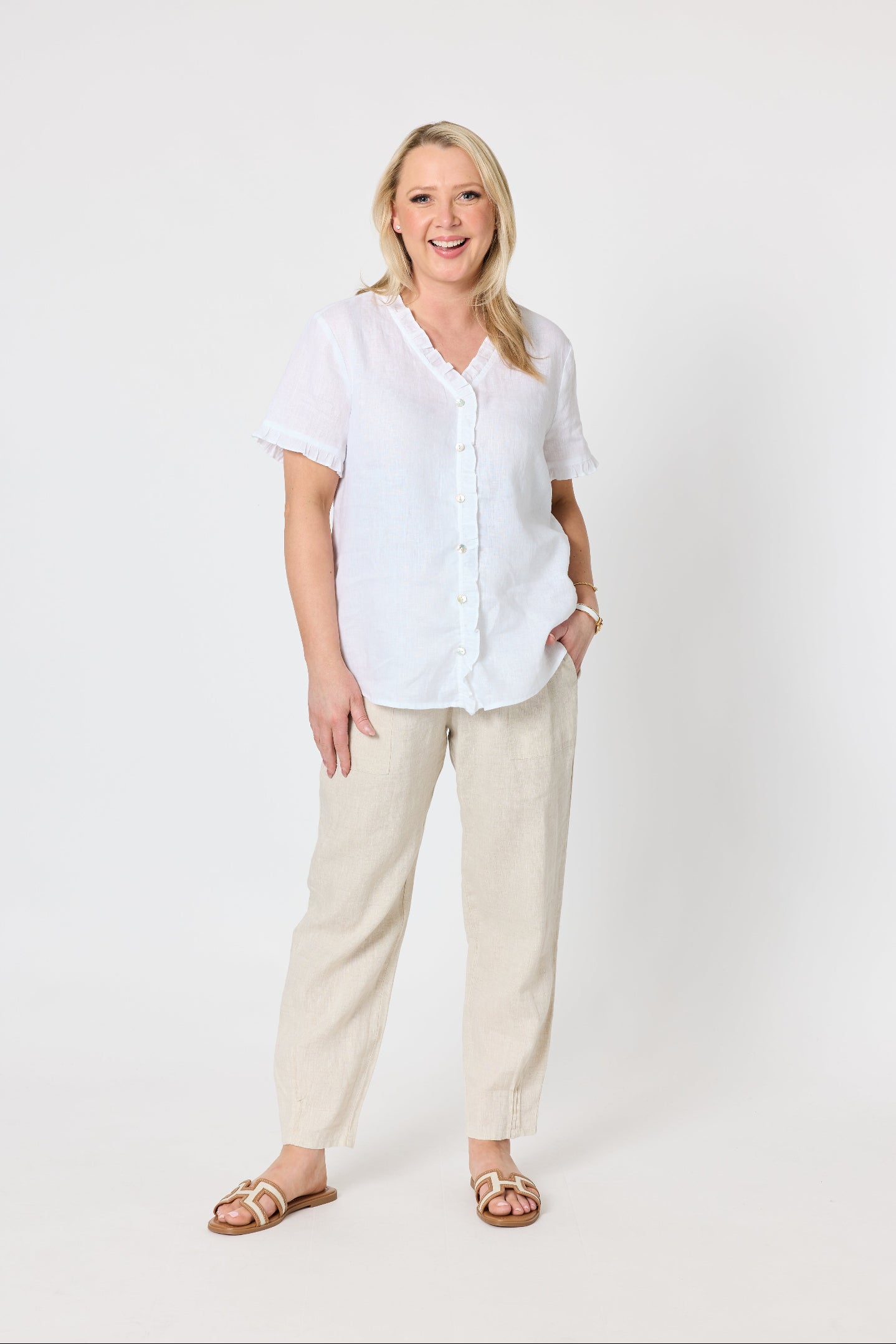 A woman modeling a white short-sleeve button-up shirt and beige linen trousers with brown sandals, standing against a plain white studio background.