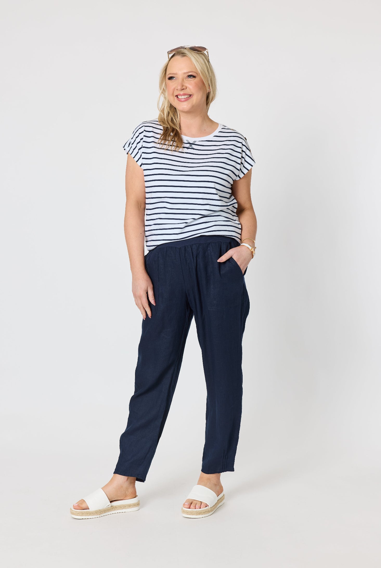A woman modeling a short-sleeve navy and white striped t-shirt paired with dark navy trousers and white slide sandals, standing against a light gray studio background.