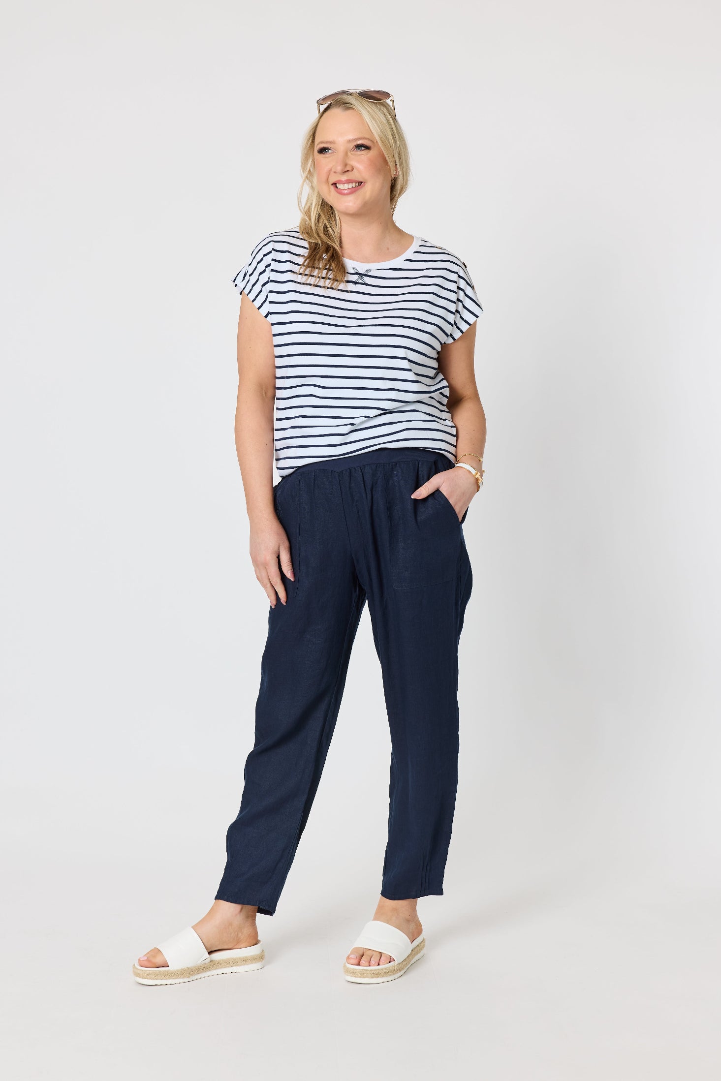 A woman modeling a short-sleeve navy and white striped t-shirt paired with dark navy trousers and white slide sandals, standing against a light gray studio background.
