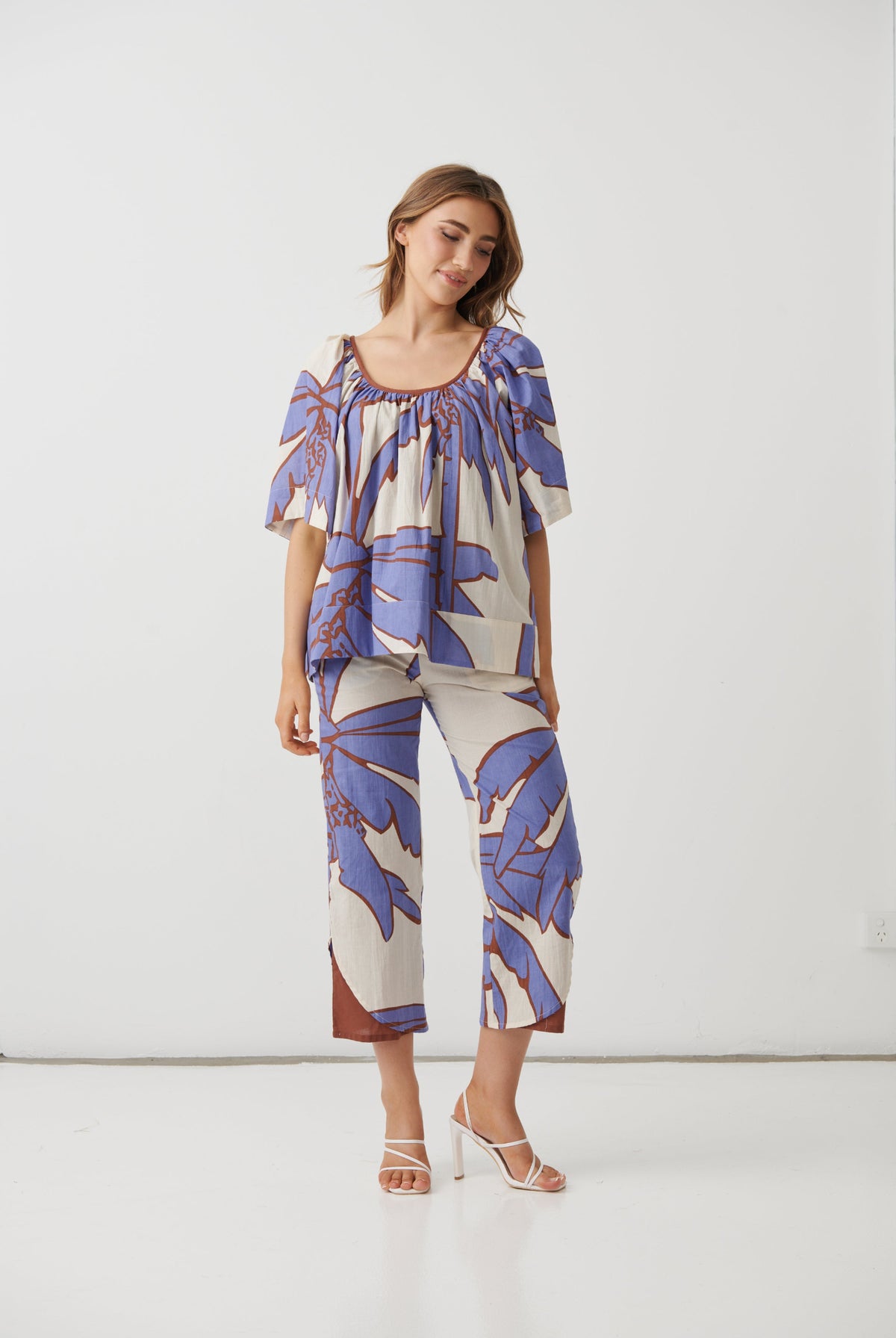 A woman modeling a matching two-piece set featuring a short-sleeve top and cropped pants with a blue and brown tropical leaf print on a cream background, set against a plain white studio background.