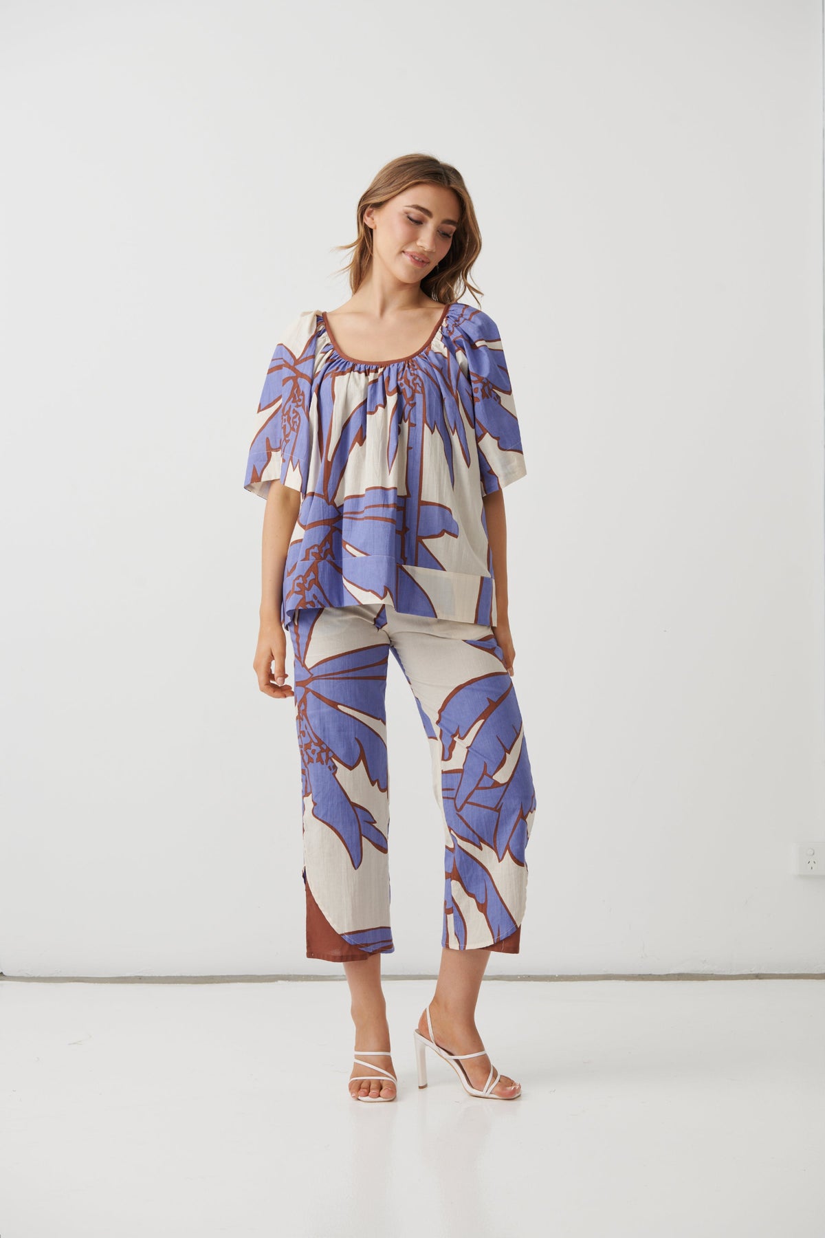 A woman modeling a matching two-piece set featuring a short-sleeve top and cropped pants with a blue and brown tropical leaf print on a cream background, set against a plain white studio background.