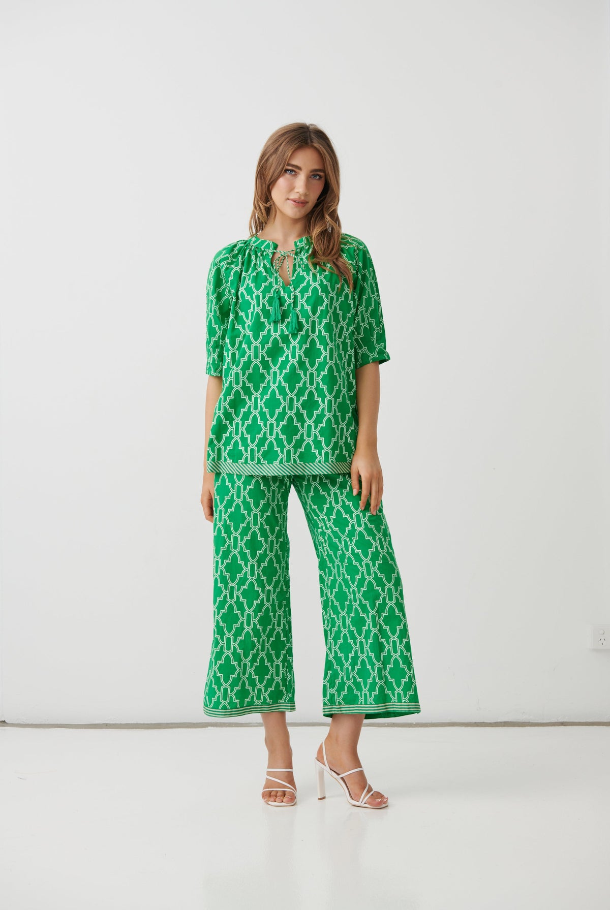 A woman modeling a green and white geometric patterned two-piece set consisting of a short-sleeve top with a keyhole neckline and matching wide-leg cropped pants, photographed against a plain white studio background.