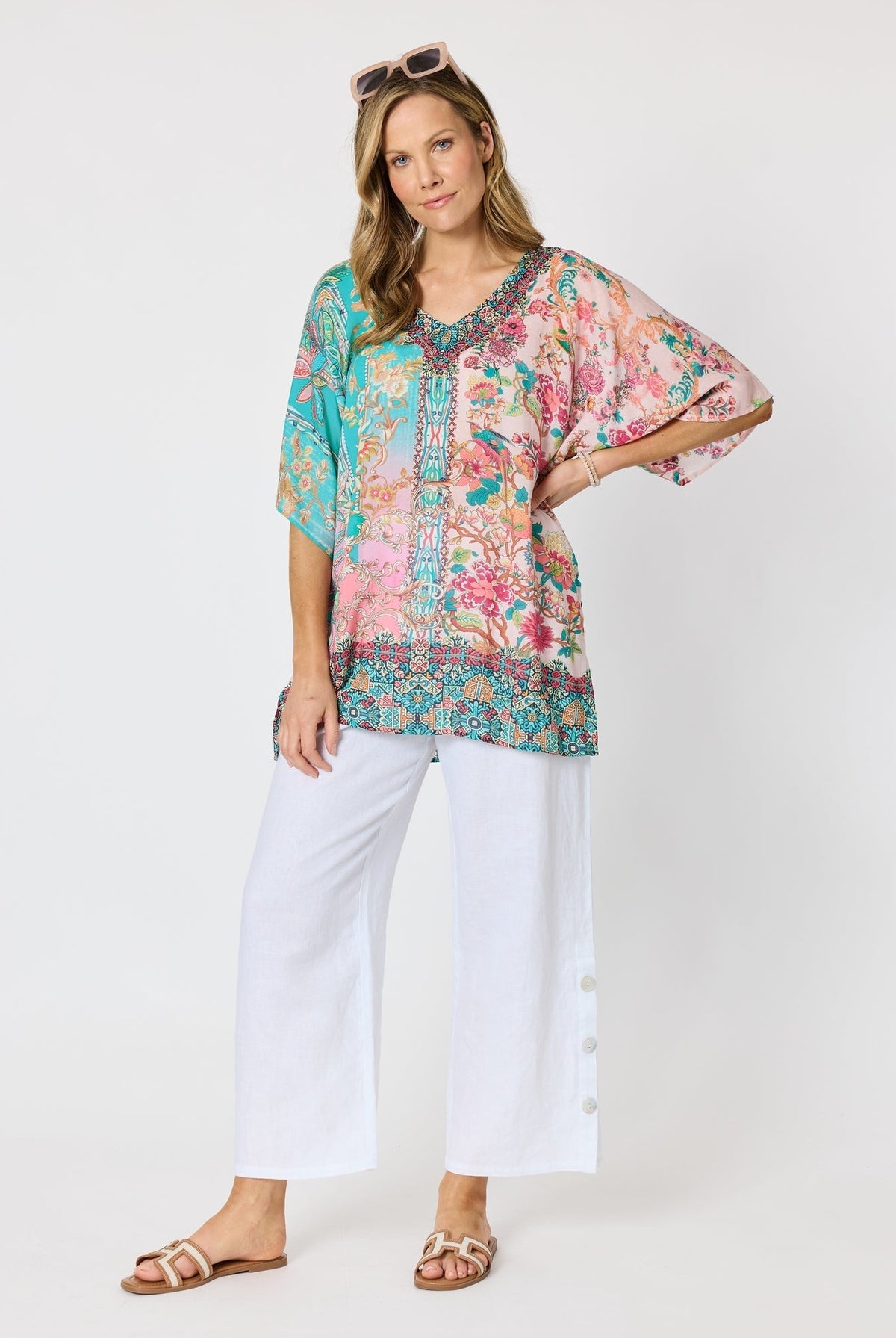 Woman wearing a colourful pink and blue printed floaty top with elbow length sleeves and detailed border and neckline and white pants against a white background.