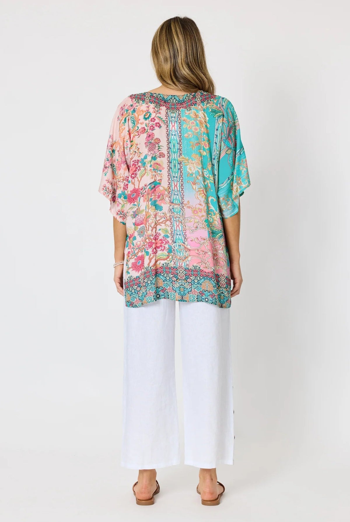 Woman wearing a colourful pink and blue printed floaty top with elbow length sleeves and detailed border and neckline and white pants against a white background.
