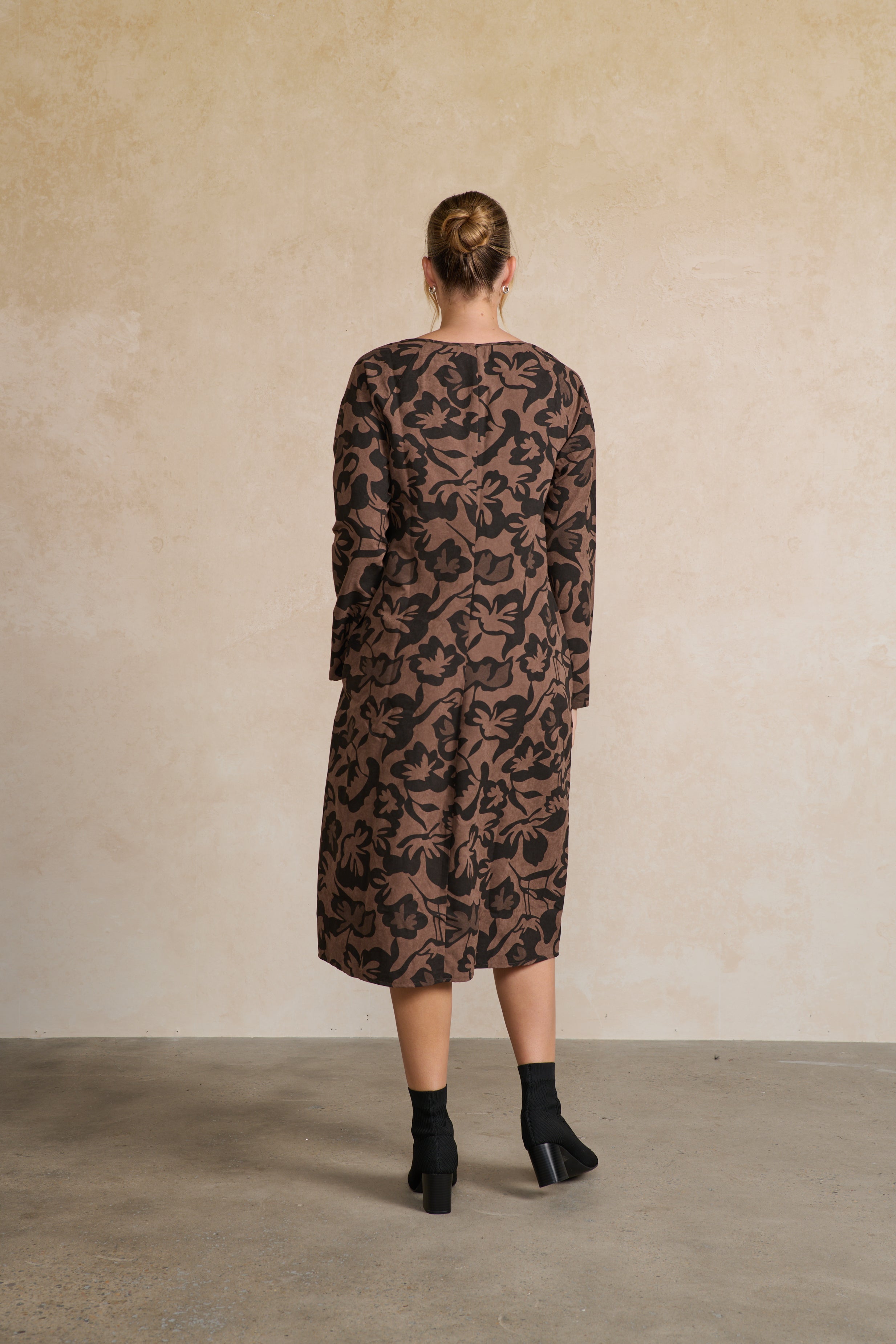 Woman wearing a brown patterned dress against a beige wall back angle