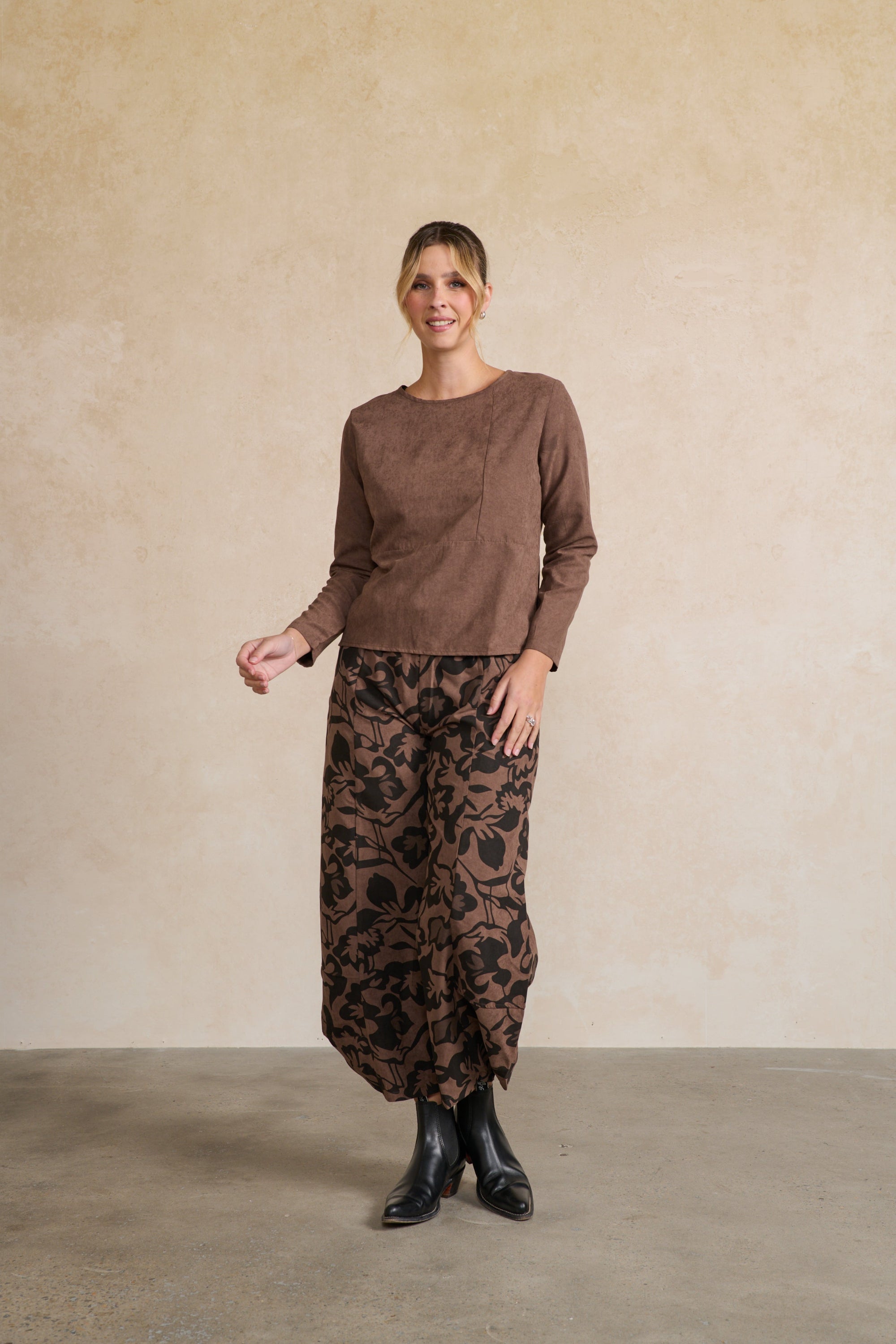 Woman wearing a brown top and patterned pants against a beige wall.