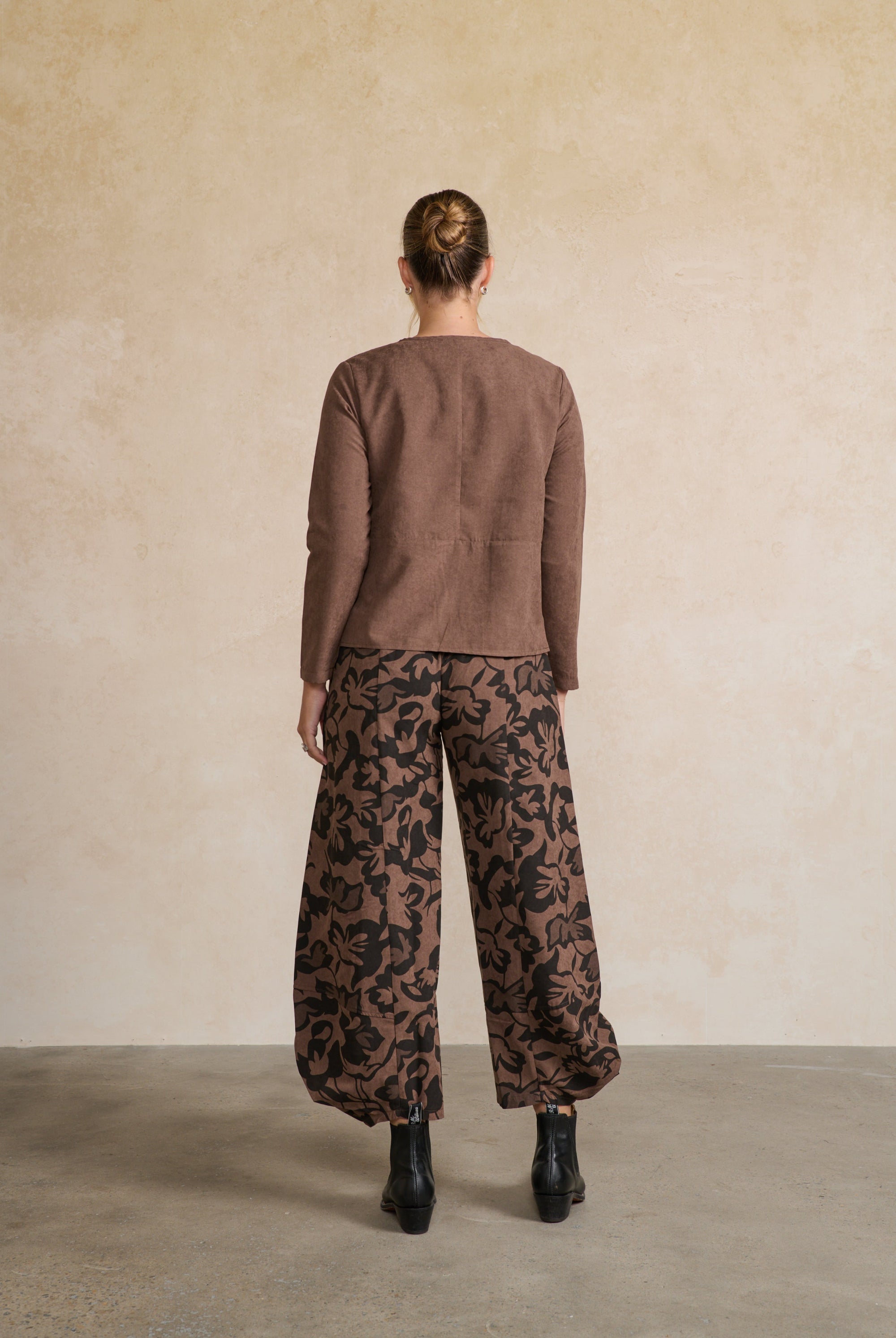 Person wearing a brown sweater and patterned pants against a beige wall back angle