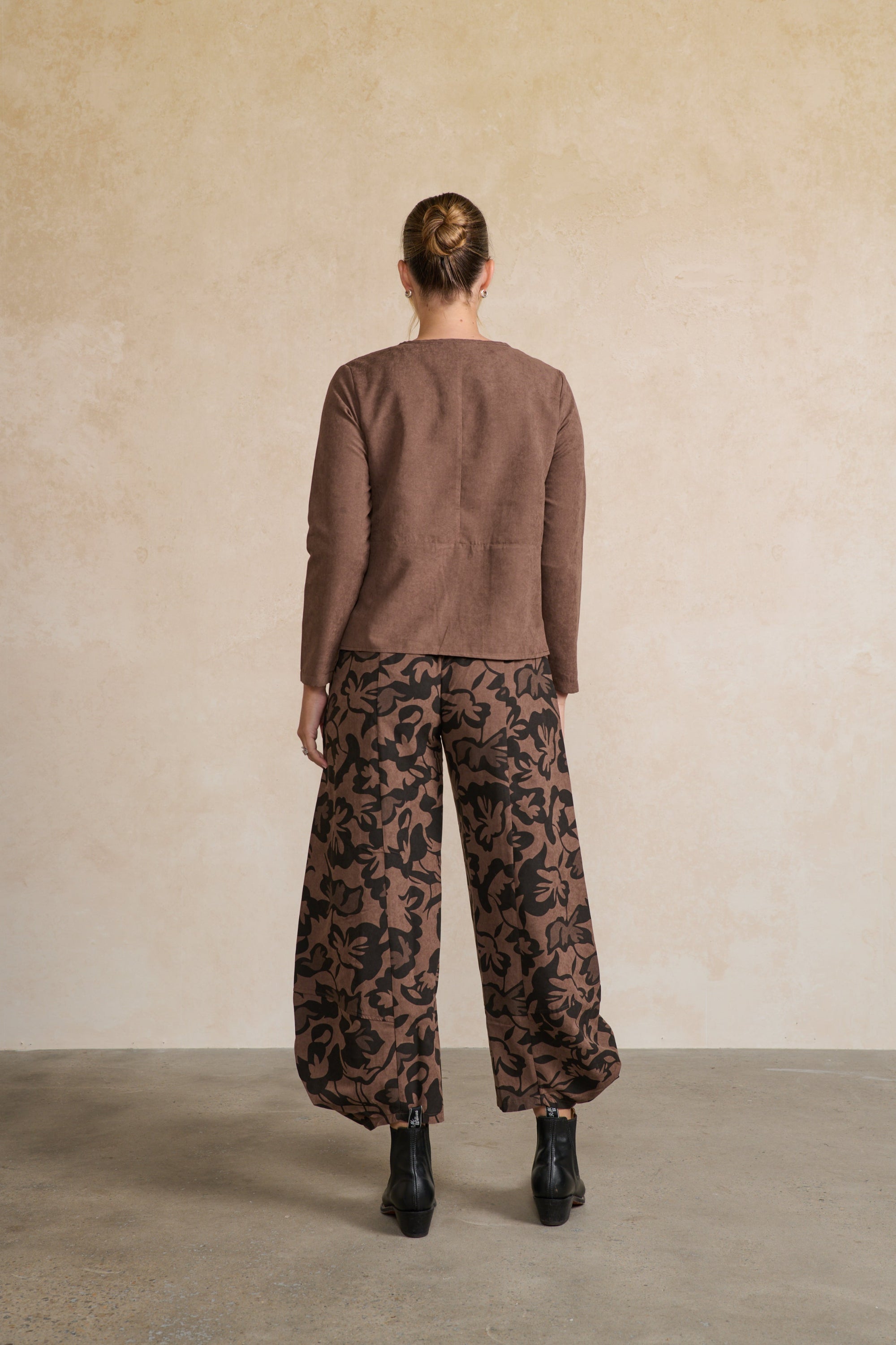 Person wearing a brown sweater and patterned pants against a beige wall back angle