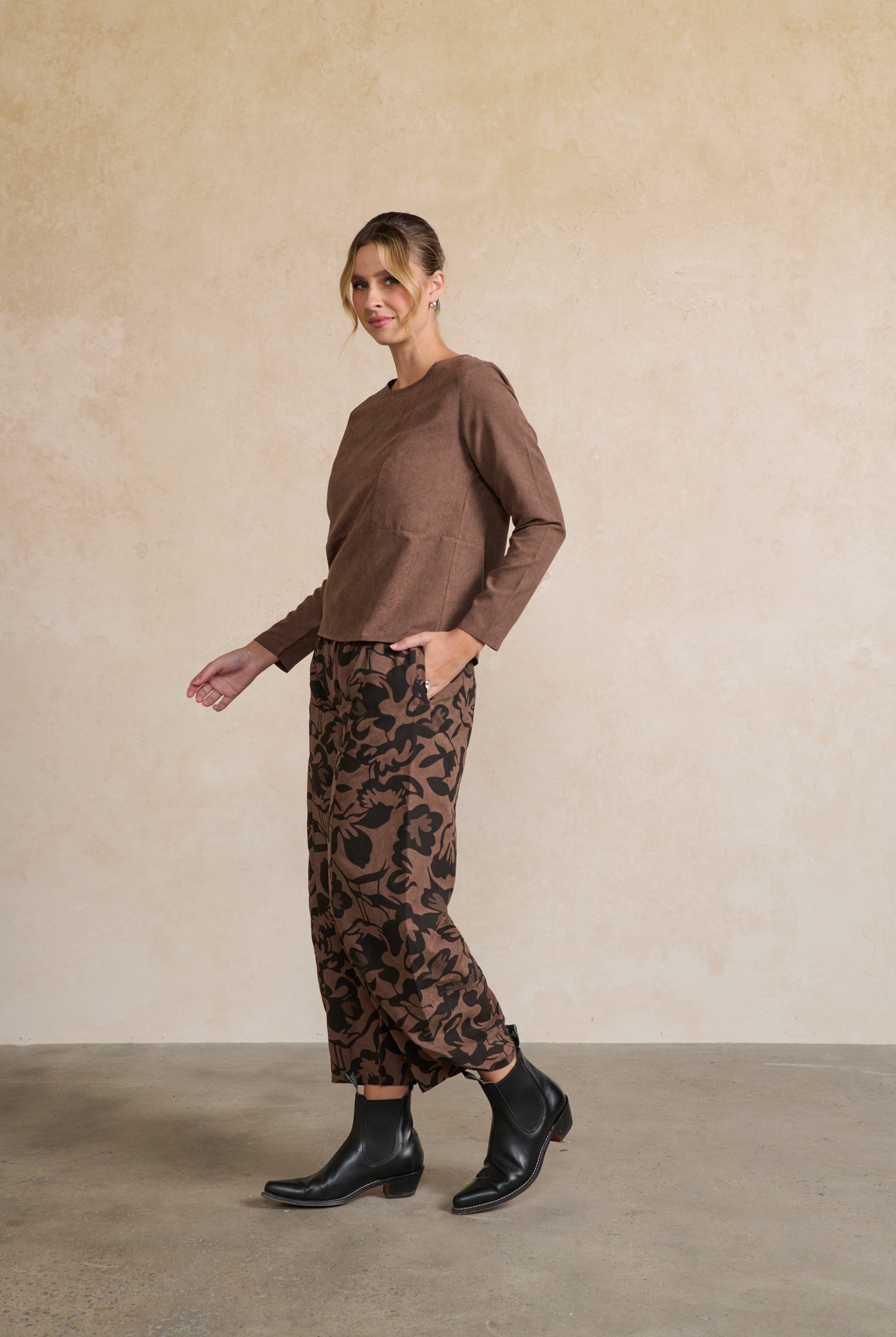 Woman wearing a brown sweater and patterned pants against a beige wall.
