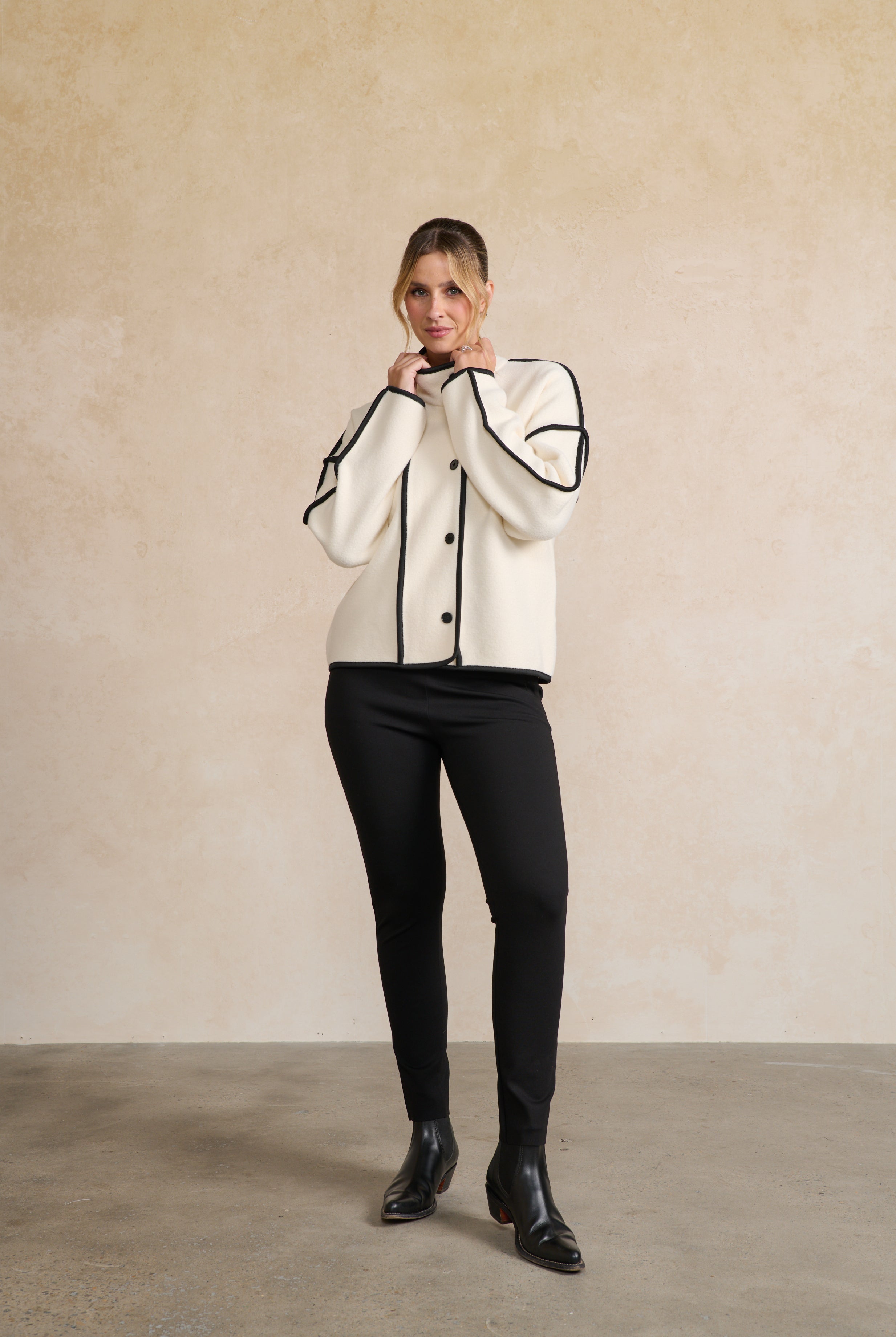 Person wearing a white jacket with black trim, black pants, and black shoes against a beige wall.