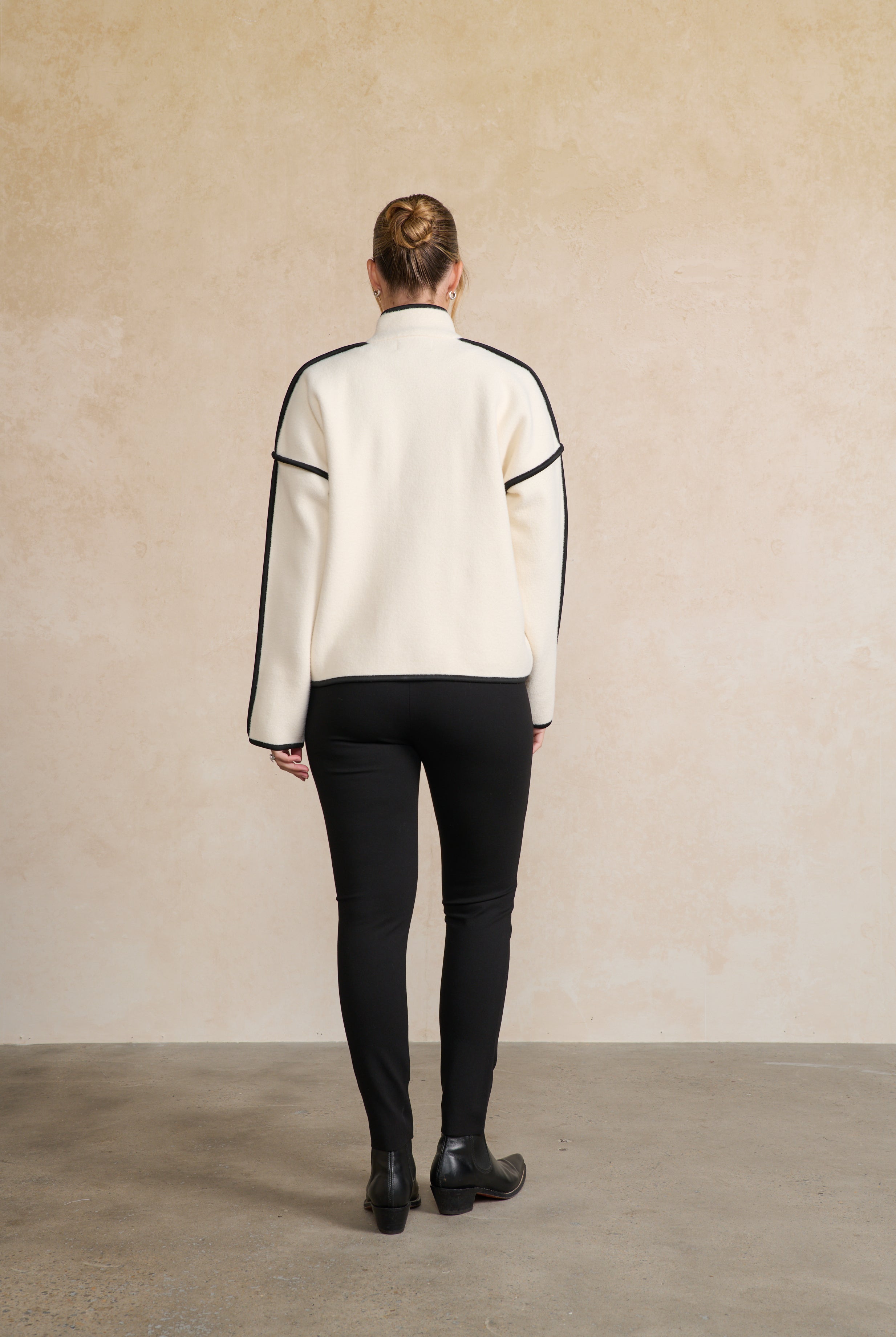Person wearing a white sweater with black accents and black pants against a beige wall back angle