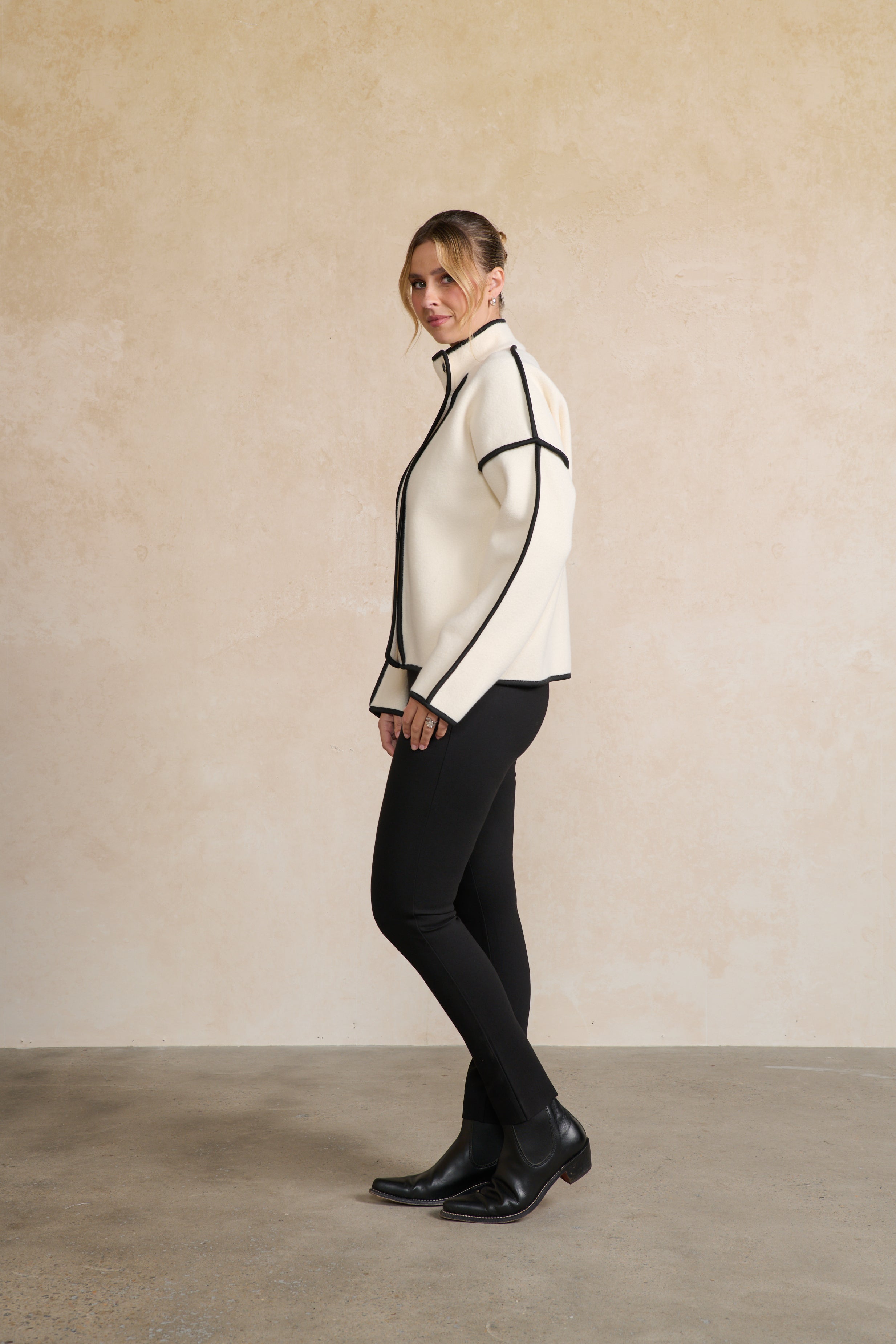 Woman wearing a white jacket with black trim against a beige wall side angle