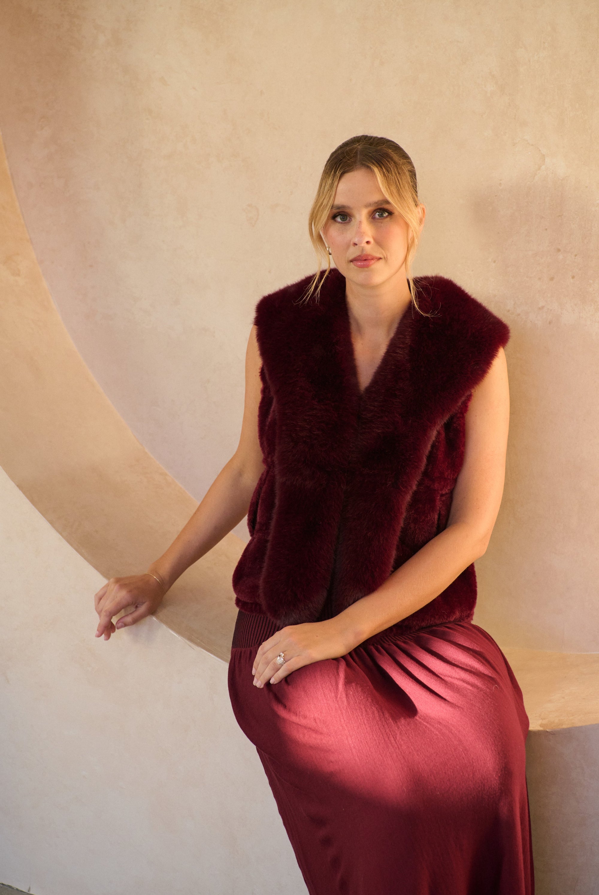 Woman wearing a burgundy dress with a fur vest against a beige wall.