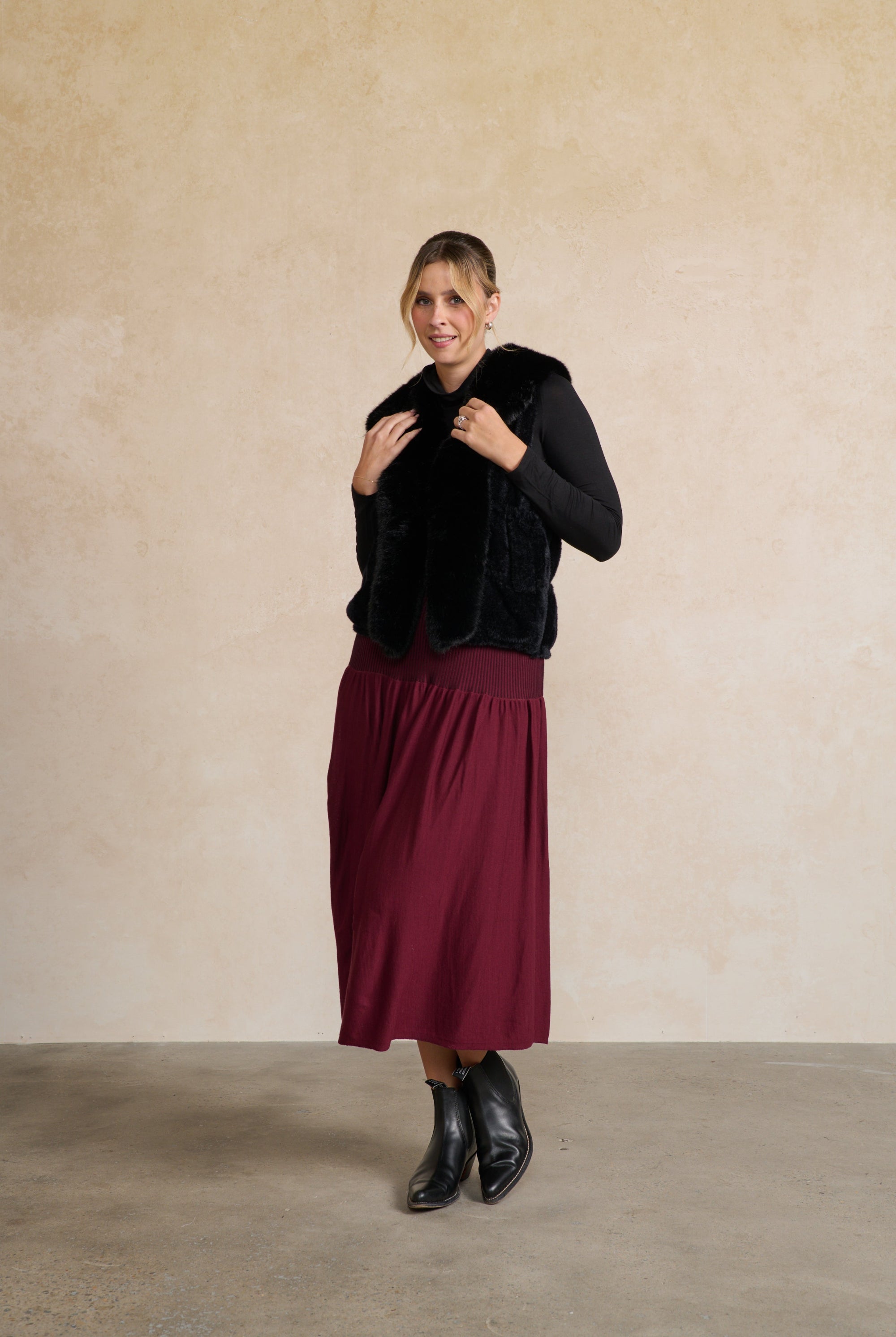 Woman wearing a black fur vest, black top, and burgundy skirt against a beige wall.