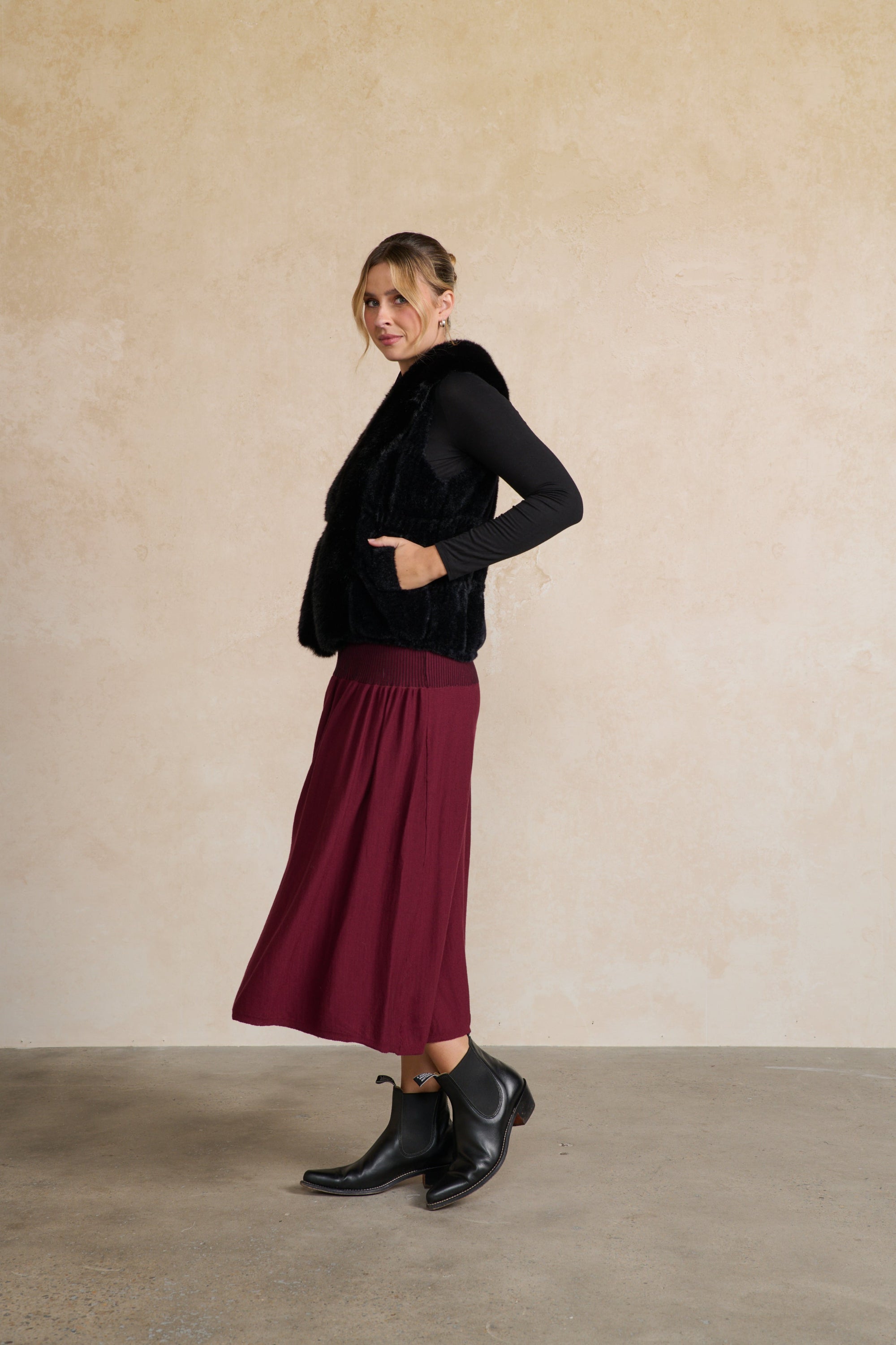 Woman wearing a blackfaux fur vest and burgundy skirt against a beige background