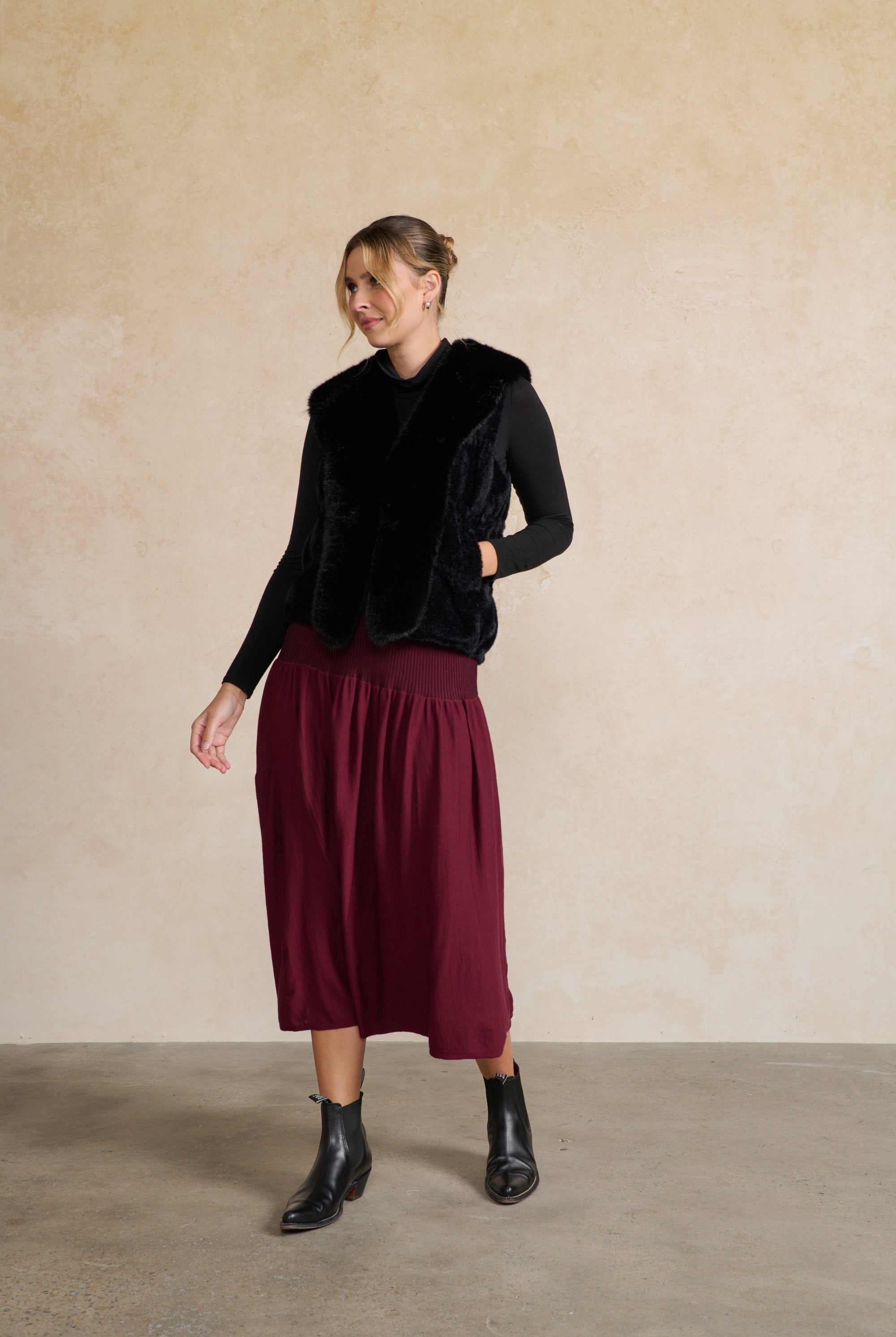 Woman wearing a black faux fur vest and burgundy skirt against a beige wall.