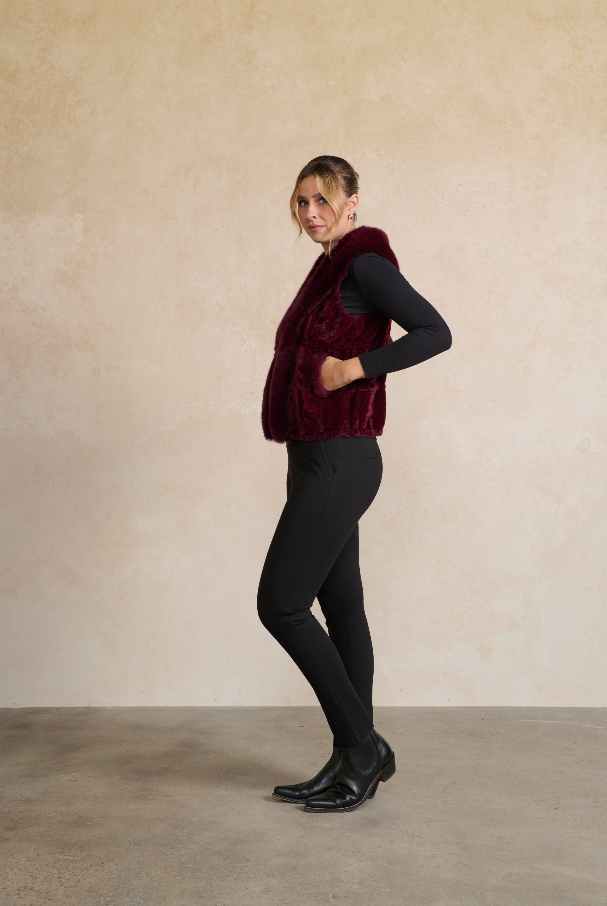 Person wearing a burgundy vest over a black top and pants against a beige wall.