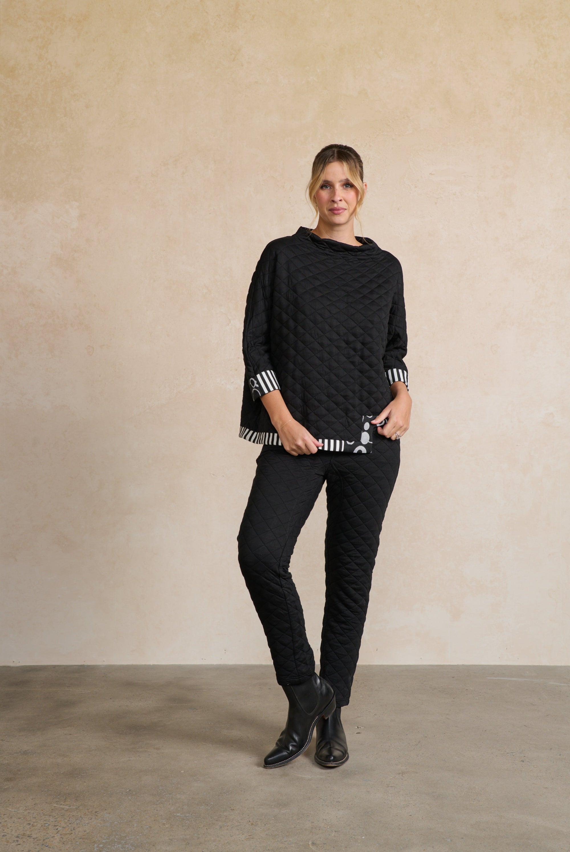 Person wearing a black quilted sweater and black pants against a beige wall.