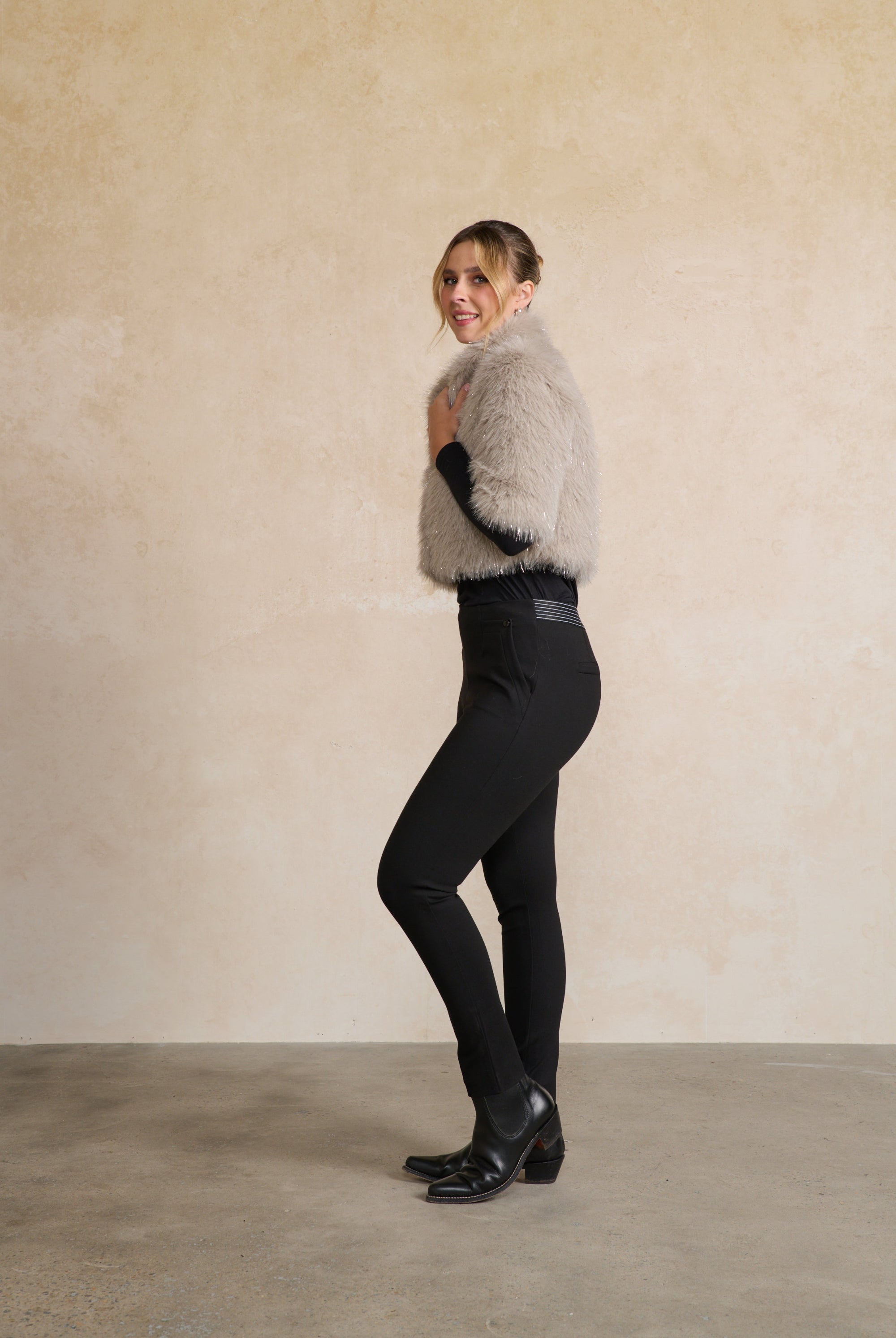 Person wearing a gray fur vest over a black outfit against a beige wall.