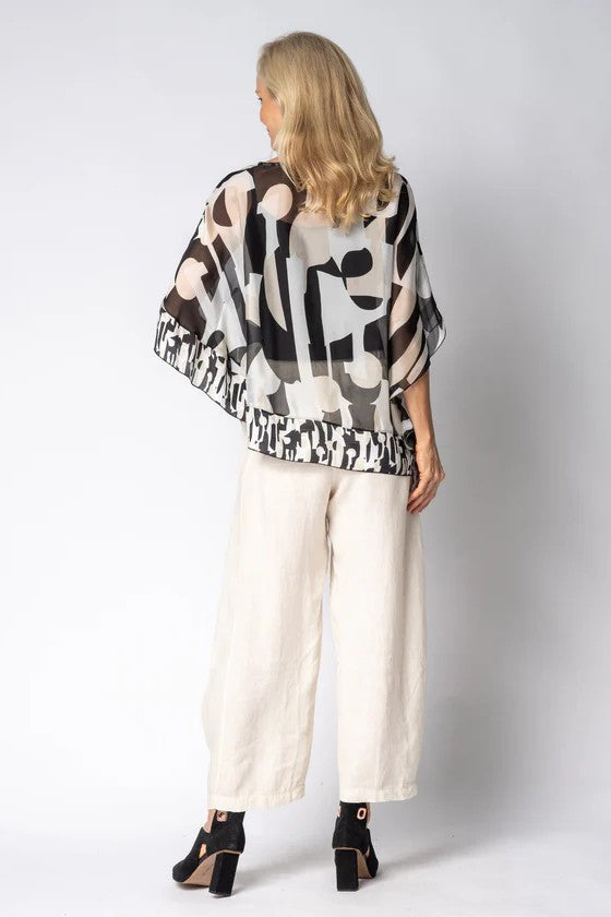 Woman wearing a black and white patterned top and white pants with a plain background back angle