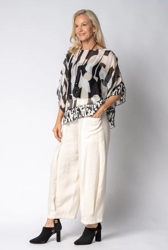 Woman wearing a patterned top and white pants against a plain background