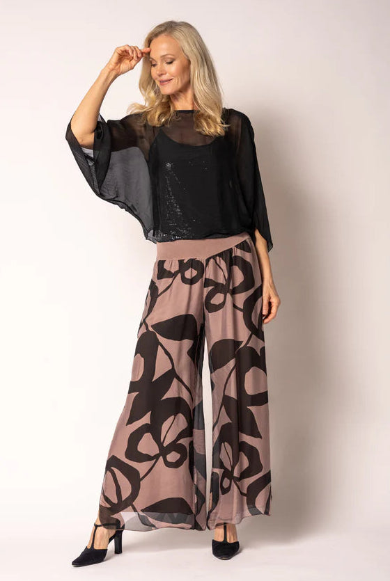 A woman modeling a sheer black mesh top with wide flutter sleeves and matching wide-leg pants featuring a black floral pattern on a dusty mauve background, paired with black heels, against a plain light background.