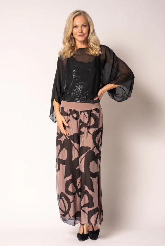 A woman modeling a two-piece outfit consisting of a sheer black long-sleeve top over a black sequined camisole, paired with a high-waisted maxi skirt in dusty pink and black abstract pattern, on a light beige studio background.