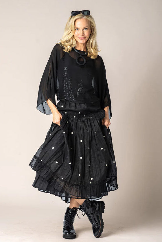 A woman modeling a black sheer long-sleeve top and a black tiered skirt with white polka dots, styled with black lace-up boots against a plain beige studio background.