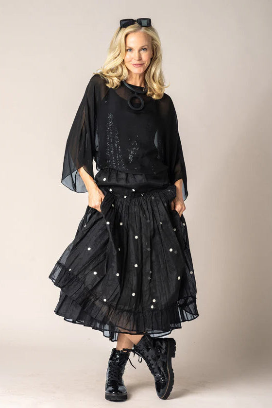 A woman modeling a black sheer long-sleeve top and a black tiered skirt with white polka dots, styled with black lace-up boots against a plain beige studio background.
