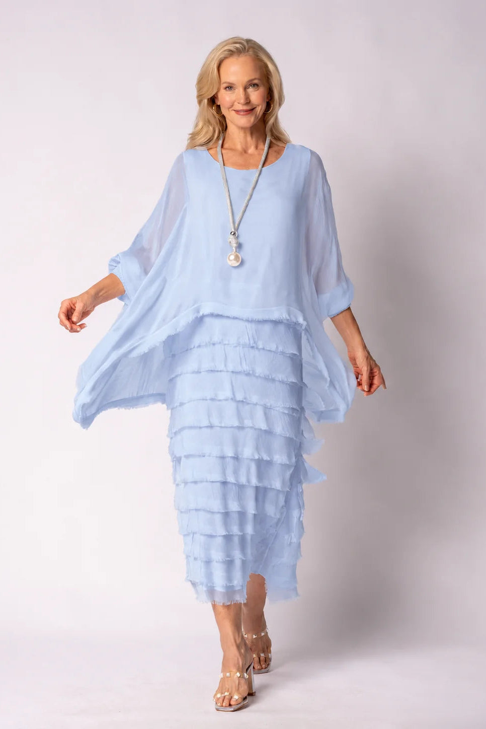 A woman modeling a light blue tiered maxi dress with a matching sheer long-sleeve top, standing against a light gray studio background.