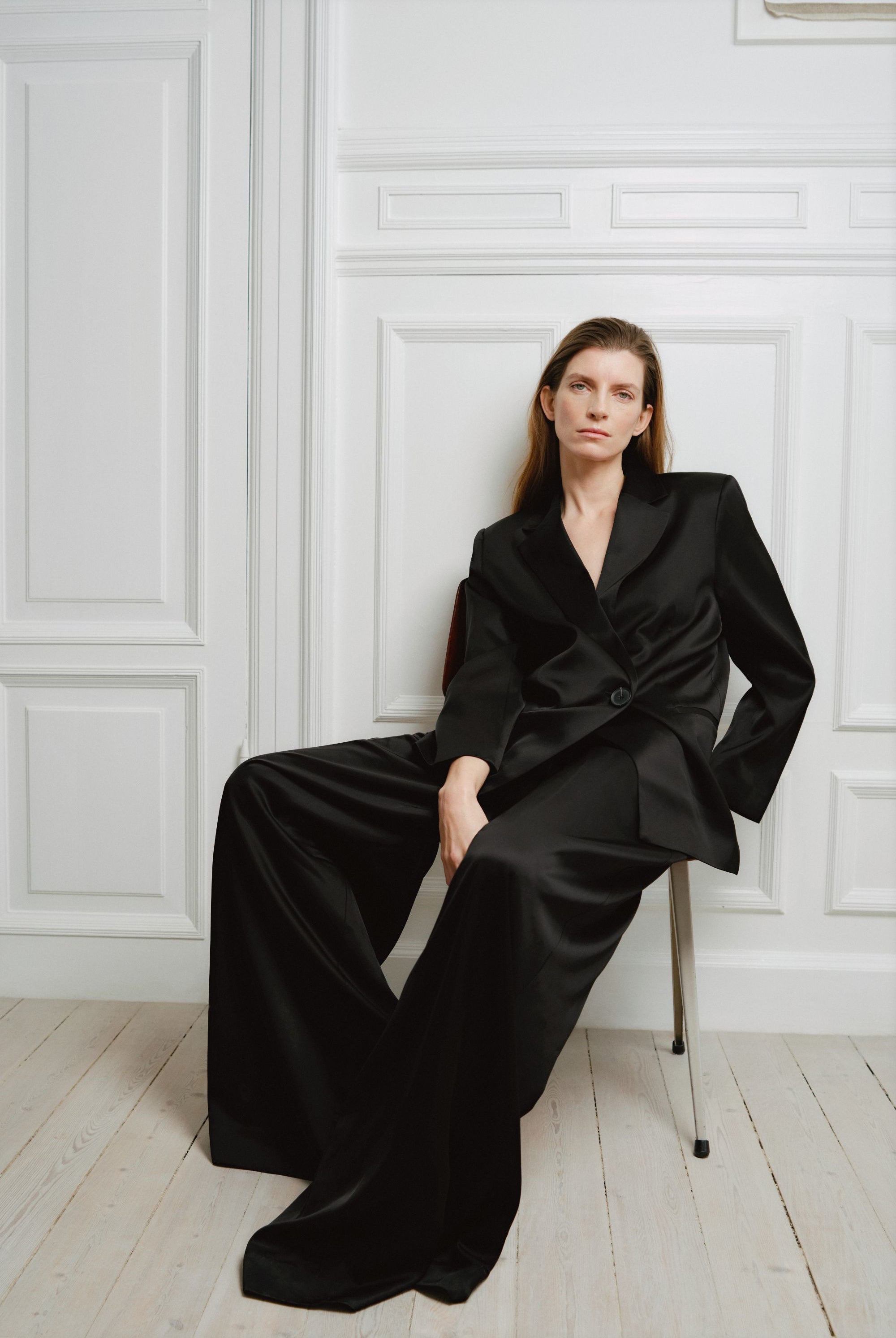 Woman wearing a black suit sitting against a white paneled wall.