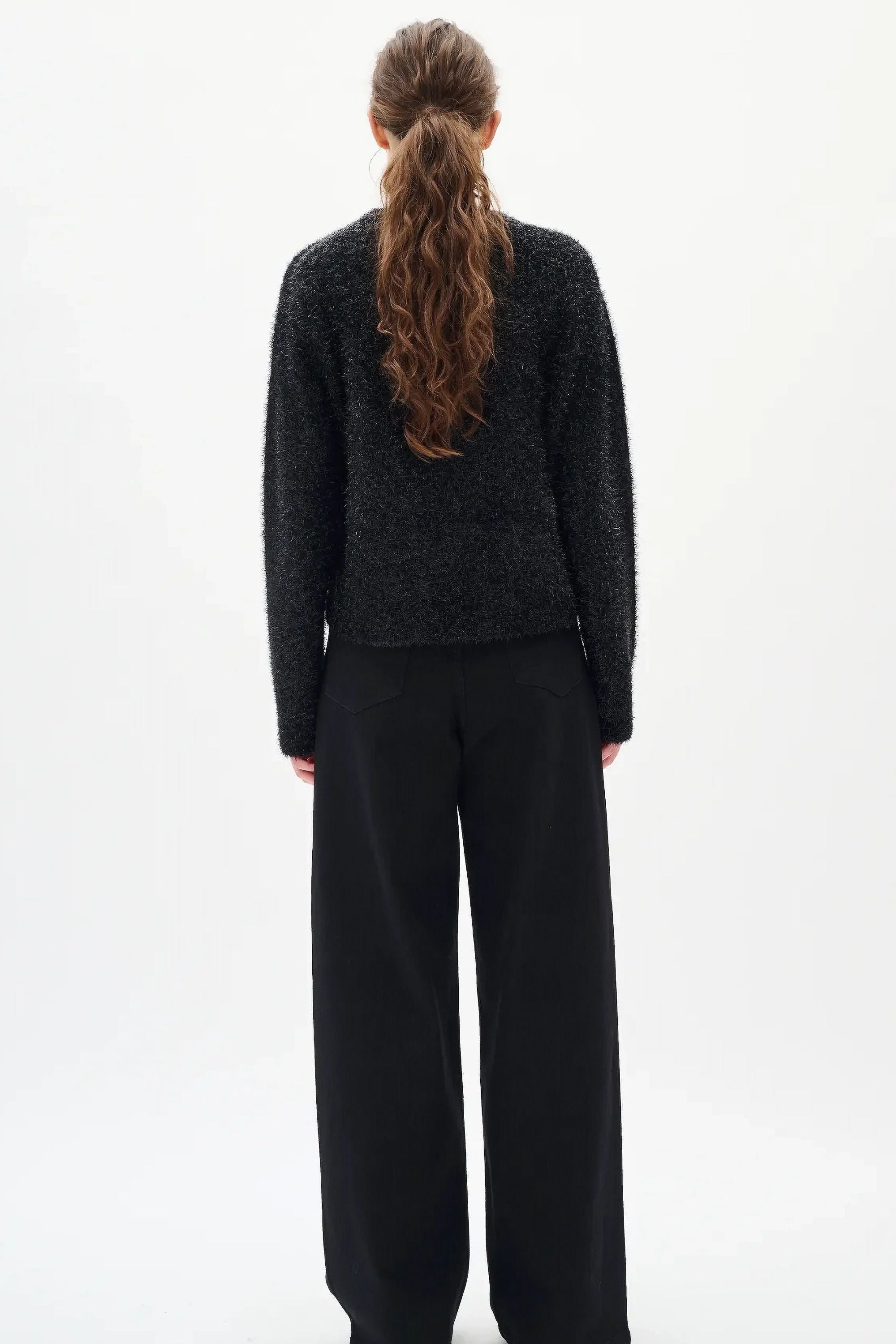Woman wearing a black textured sweater and diamante black jeans on a white background back angle